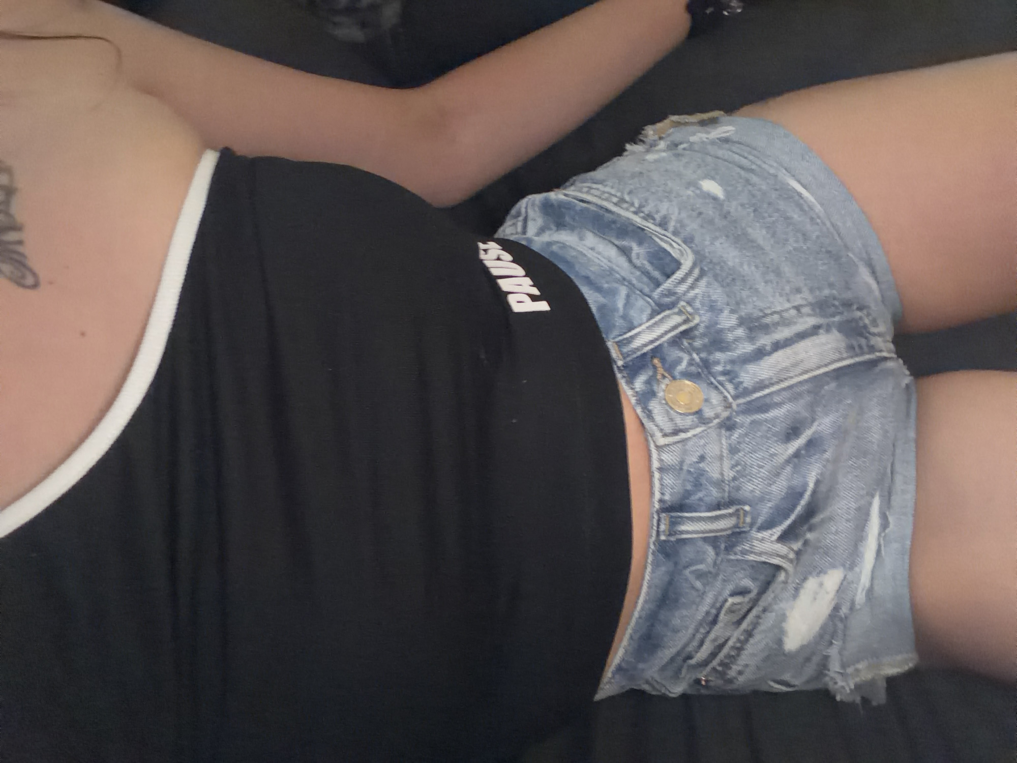 A person is wearing a black, one-shoulder top with white trim and the word "PAUSE" in white lettering. They are also wearing light-wash, distressed denim shorts. The image is a close-up shot, focusing on the person's torso and upper legs. The background is dark and out of focus, suggesting an indoor setting. There are no discernible elements that would indicate a specific location within Plovdiv, Bulgaria.