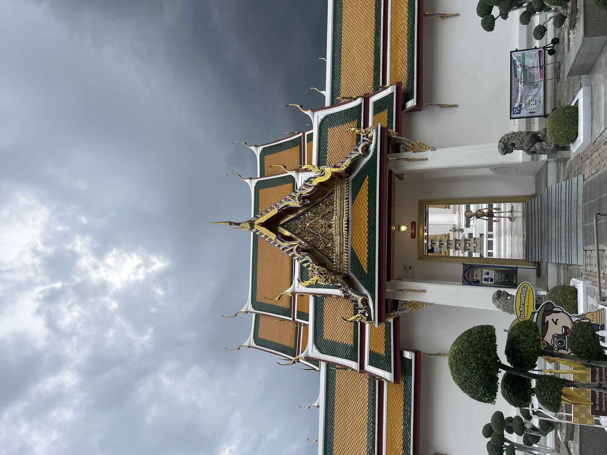 The image depicts the entrance to a temple in Bangkok, Thailand, under a cloudy, overcast sky. The temple's roof features a traditional architectural style with orange and green tiles, adorned with ornate golden finials. In front of the temple, a white wall serves as a backdrop, with a doorway leading to an interior courtyard. The foreground is decorated with meticulously trimmed topiaries and a statue of a cartoon character holding a camera. To the right of the entrance, a stone lion statue guards the pathway, and a sign provides information about parking and services. Inside the doorway, a slender, mannequin-like figure stands in the courtyard. The overall atmosphere suggests a serene and culturally rich setting, possibly Wat Suthat or a similar temple within Bangkok.