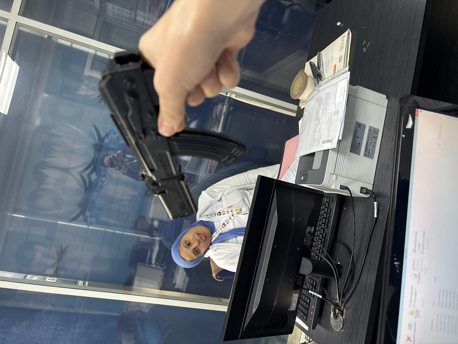 The image captures an indoor scene within a modern office environment, located in Meknes, Morocco. In the immediate foreground, a light-skinned hand holds a black replica assault rifle, resembling an AK-47, with its muzzle pointed towards the mid-ground. The firearm appears to be a prop weapon, featuring an inserted magazine.
Directly behind the hand, a dark-surfaced office desk is visible, equipped with a black computer monitor and a black keyboard. To the right of these items sits a light gray HP LaserJet M1536dnf MFP printer/scanner, with several documents stacked on its top surface, including an official-looking envelope. A red folder and a roll of tape are also present on the desk.
Separating the foreground desk from the mid-ground is a clear glass partition adorned with blue, stylized wave-like or floral patterns. Behind this partition, an adult female is seated in a blue office chair at another dark desk. She wears a light blue hijab and a white top, with an identification lanyard displaying multiple small, colorful cartoonish images around her neck. Her head is tilted, and she is seen with her tongue playfully extended, indicating a non-serious or humorous interaction with the person holding the replica weapon.
Reflections of the office lighting and the individual taking the photograph (a female holding a smartphone) are visible on the glass partition. The background, seen through the partition, suggests additional office space.