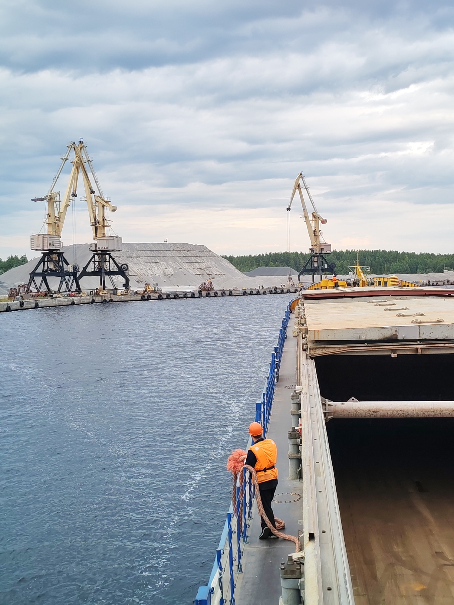 A worker in an orange safety vest and hard hat stands on the deck of a large cargo ship, holding a thick rope. The ship is docked at a port with a long pier. On the pier, two large yellow cranes are positioned next to enormous piles of light-colored gravel or sand. Behind the piles of aggregate, a dense green forest borders the port. In the distance, another, slightly smaller crane is visible. The sky above is filled with dramatic, cloudy formations. The water is dark blue and appears choppy. The scene suggests industrial activity, likely related to bulk material handling at a port. The worker's stance and the rope imply preparation for or completion of docking or loading/unloading operations.