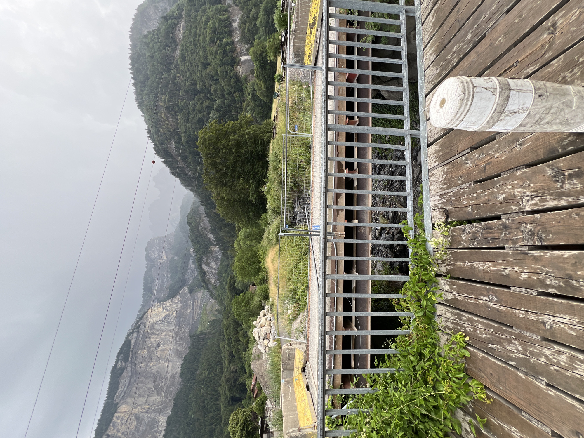 A wooden walkway leads to a metal railing overlooking a bridge under construction. The bridge has a concrete deck with yellow barriers and a metal substructure. Beyond the bridge, a steep, rocky mountain with a forested slope rises towards a cloudy sky. Electrical wires are visible in the sky. In the mid-ground, there are piles of rocks and construction debris, as well as some buildings. The scene is overcast and appears to be in a mountainous region, possibly near Sallanches, France.