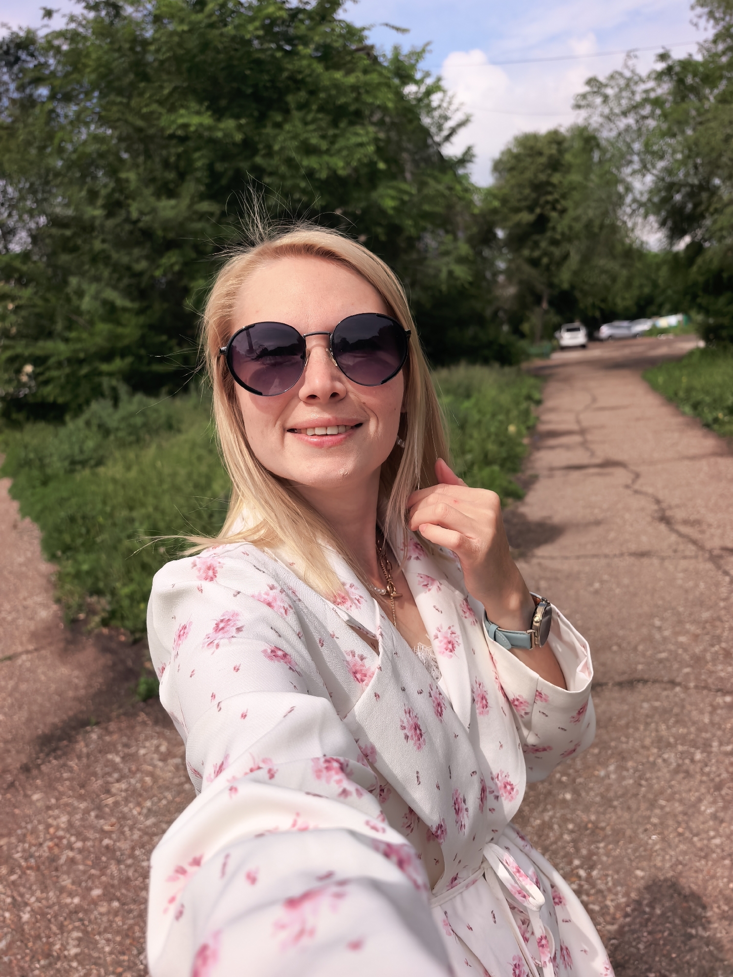 A blonde woman, appearing happy and relaxed, is captured in a selfie outdoors in Sterlitamak, Russia. She wears large, round, dark sunglasses and a white wrap dress adorned with delicate pink floral patterns. A light blue watch with a gold face is visible on her left wrist, and a necklace with a pearl-like pendant adorns her neck. Her hair is slightly windswept.
Behind her, a reddish-brown paved path, showing some cracks, splits into two directions, lined by lush green trees and bushes on both sides. The setting appears to be a park or a green recreational area. In the distant background, several cars are visible along the path under a blue sky with scattered white clouds. The bright, natural lighting indicates it is daytime, likely a sunny or partly cloudy day. There is no visible text in the image.