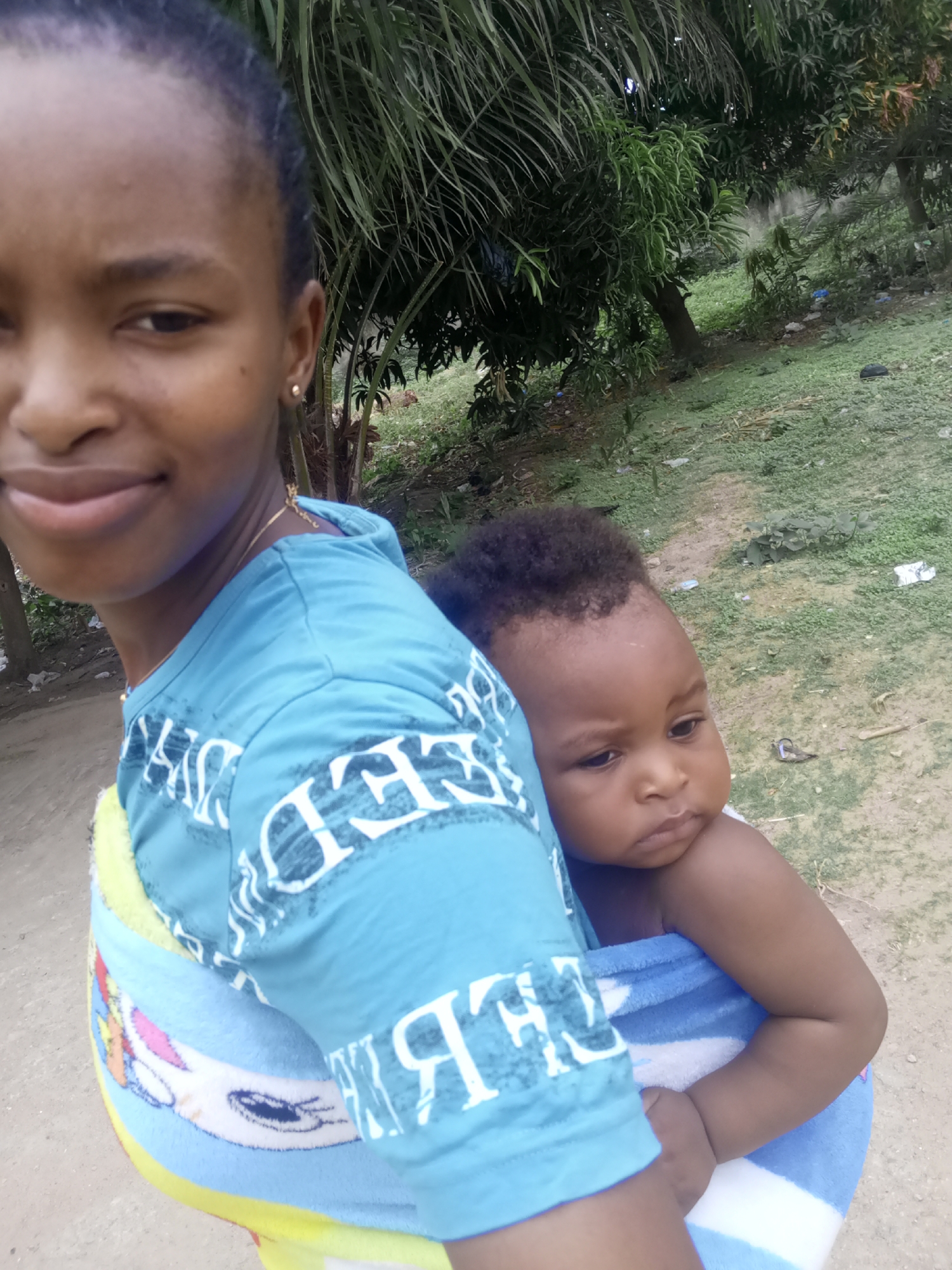 In the city of Unknown, Unknown, a woman is carrying a baby on her back. The woman is wearing a light blue t-shirt with white writing on it. The baby is wrapped in a blue and white striped towel. The background includes trees, grass, and some litter. The ground appears to be a mix of dirt and grass.