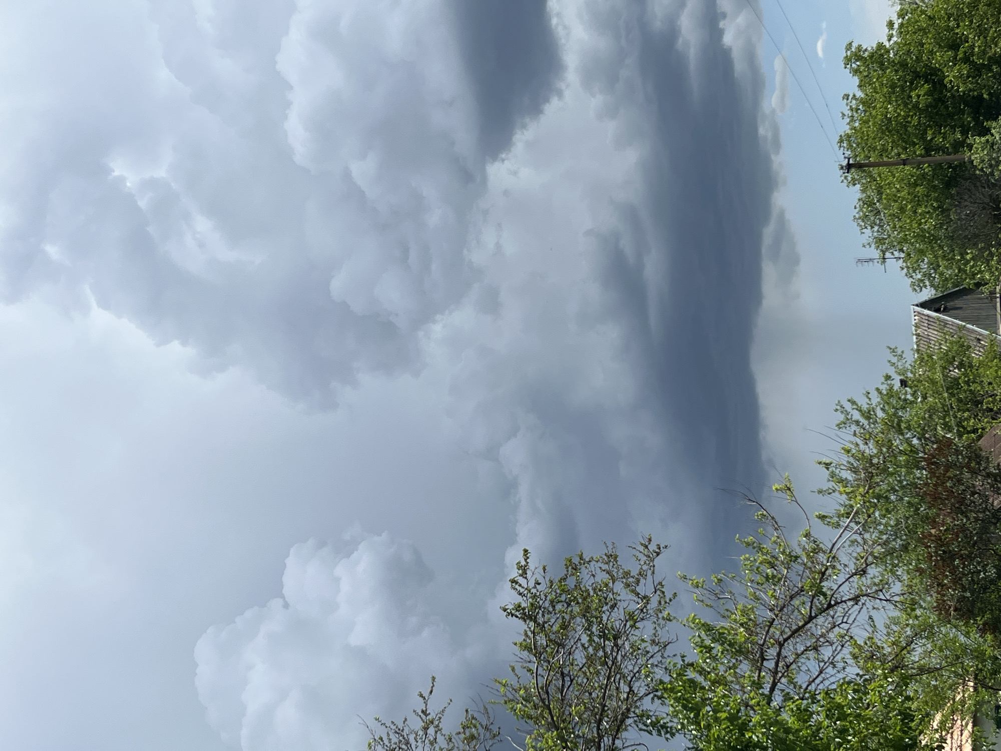 The image captures a dramatic sky with ominous, dark storm clouds dominating the scene. In the foreground, there are trees with green leaves, partially obscuring the view. A utility pole with wires is also visible, along with the roof of a building. The location is likely Poltavka, Ukraine, though no explicit details confirm this location. There are no people visible in the image, and the primary subject is the approaching storm.