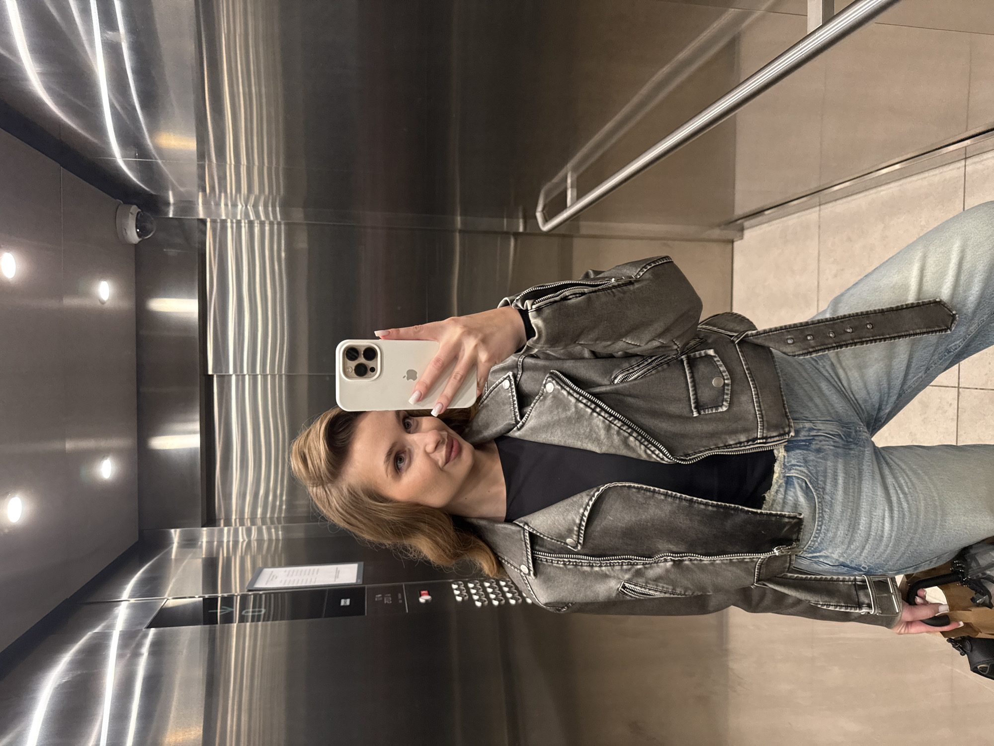 A woman is taking a selfie in an elevator. The elevator has a shiny metal interior, with a handrail visible along the top right. There is a security camera in the upper-left corner. The woman is wearing a grey belted leather jacket, a black top, and blue jeans. She is holding a white iPhone to take the selfie. The button panel of the elevator is visible to her left. There is no information to confirm that the scene is set in Belgrade, Serbia.