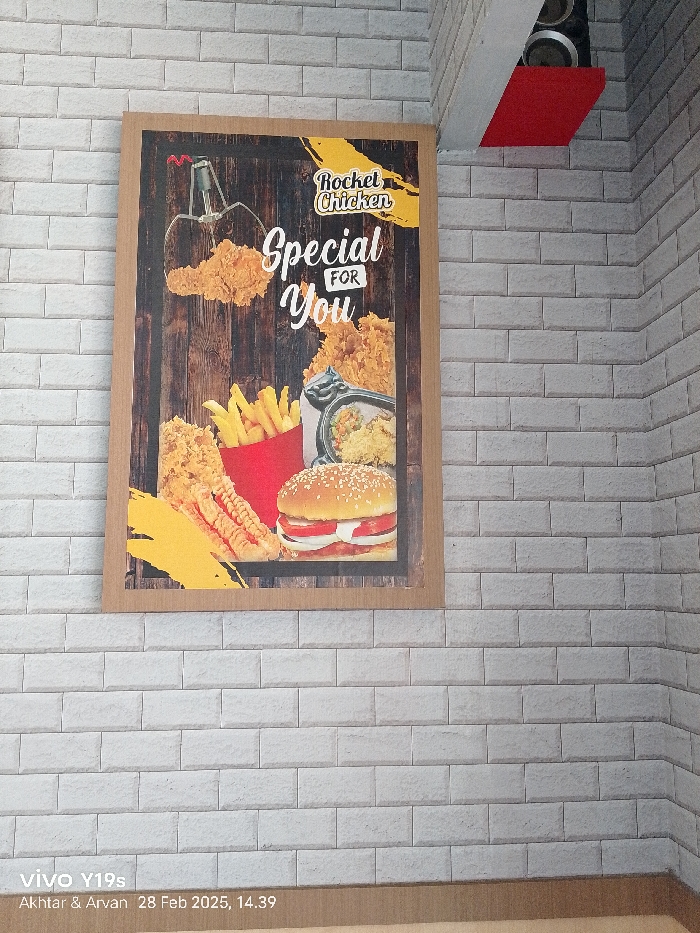 A poster advertising "Rocket Chicken" with the slogan "Special For You" is mounted on a white brick wall. The poster depicts various fried chicken items including drumsticks and wings, a hamburger, french fries, and a side dish. The poster is mounted in the corner of the wall. The coordinates -6.318, 107.986, locate the scene in the city of Patrol, Indonesia.