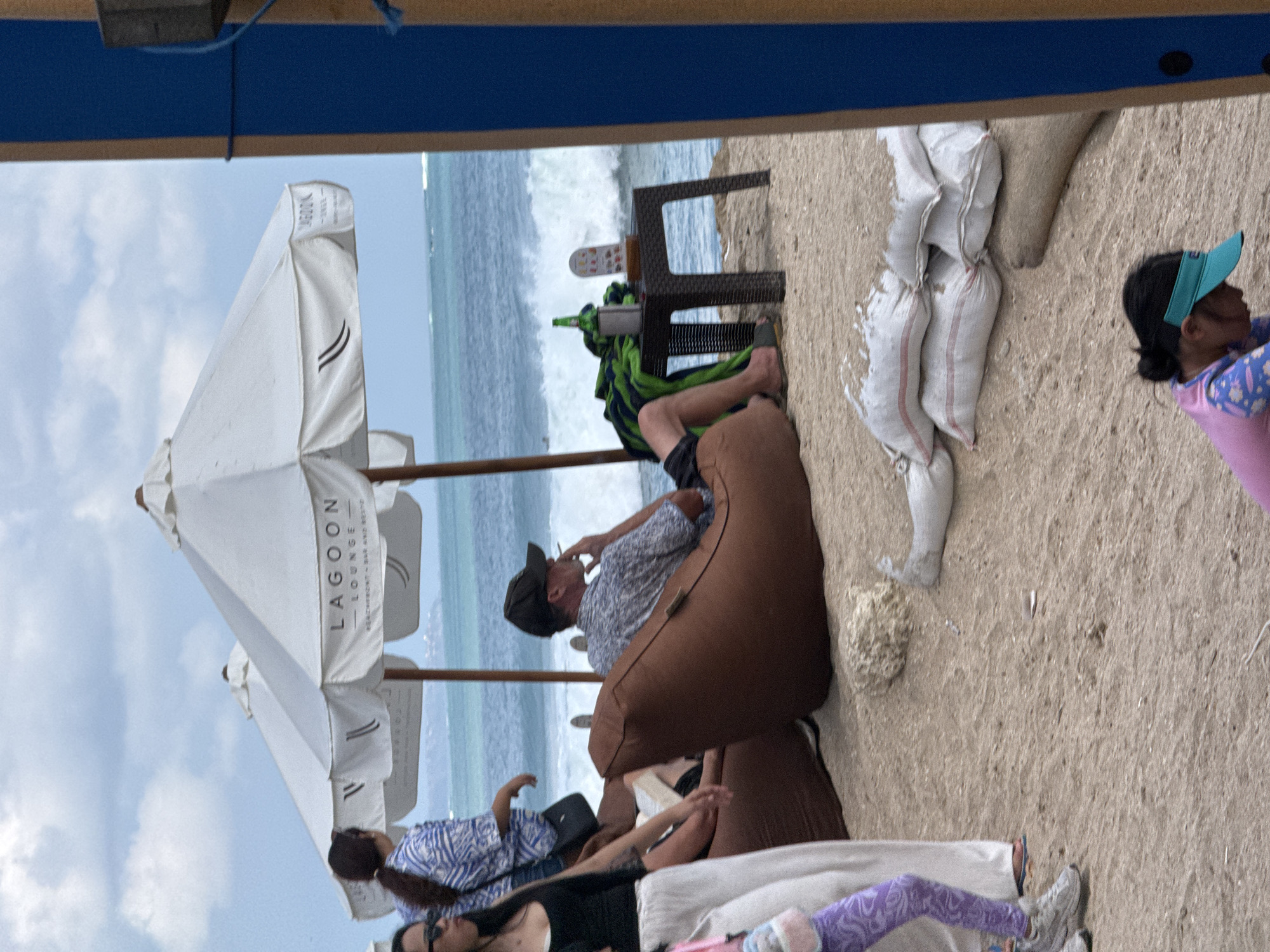 This image depicts a relaxed scene at a beachfront lounge in Kuta, Indonesia. Several people are seen lounging on beanbag chairs and lying on the sand. A man wearing a patterned shirt and a baseball cap is seated on a brown beanbag chair, seemingly smoking a cigarette. In the background, the ocean with waves crashing onto the shore is visible. White umbrellas, one of which has the text "LAGOON LOUNGE - BEACHFRONT - BAR AND RESTO" printed on it, provide shade. Several white sandbags are piled on the beach, possibly for protection or as seating. A woman wearing a blue visor and a colorful shirt is also visible further down the beach. The sky is cloudy, suggesting it might be a partly cloudy day. The overall atmosphere appears to be one of leisure and relaxation at a tropical beach resort.