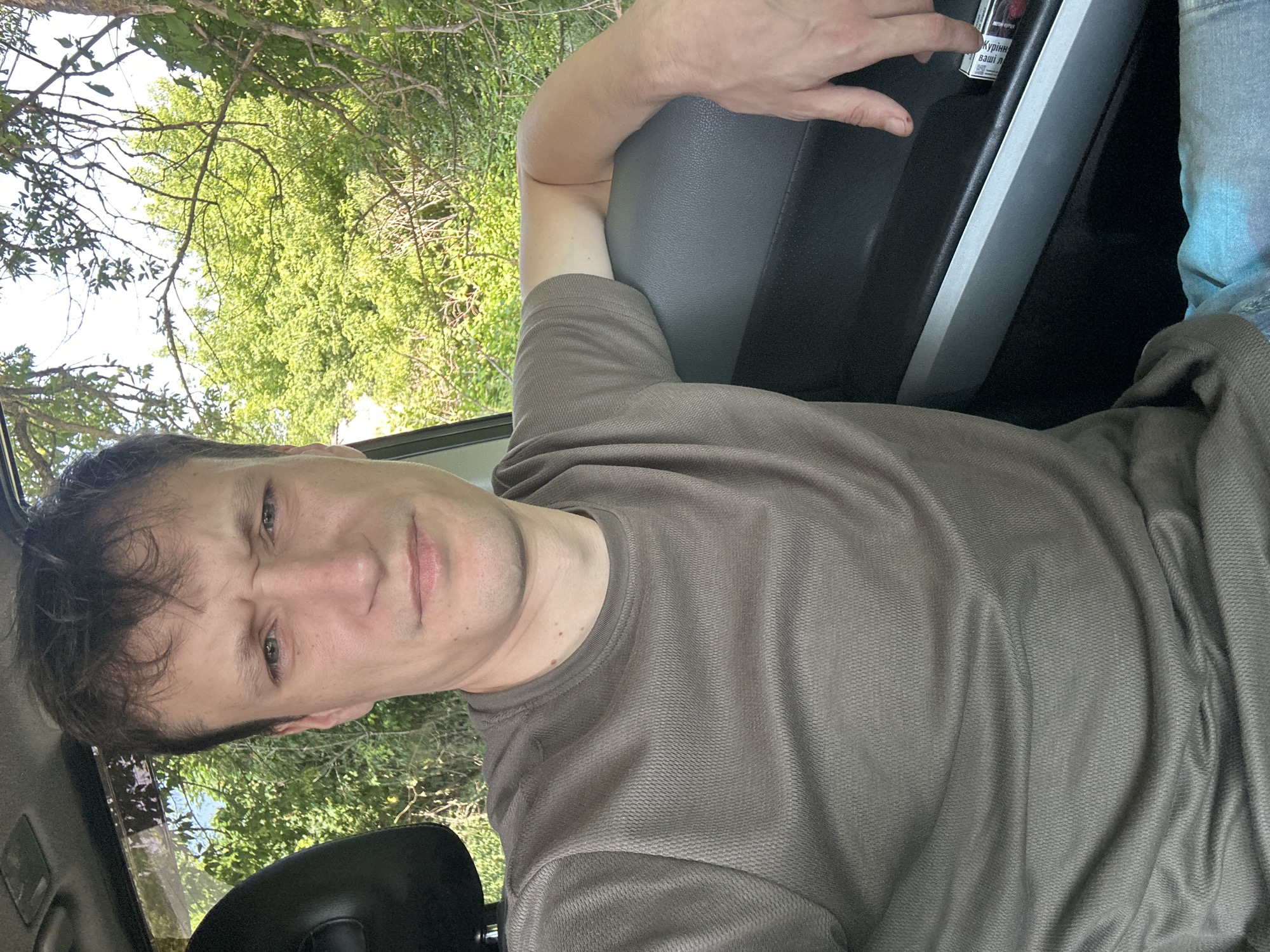 A man is sitting inside a vehicle, facing the camera and looking off to his left with a strained expression. He has dark, disheveled hair and is wearing a dark grey t-shirt. The interior of the vehicle is visible, including the car door, and the exterior shows green trees and branches through the window, suggesting a rural or natural setting. A pack of cigarettes is on the car door armrest. The location is noted as Makiv, Ukraine.