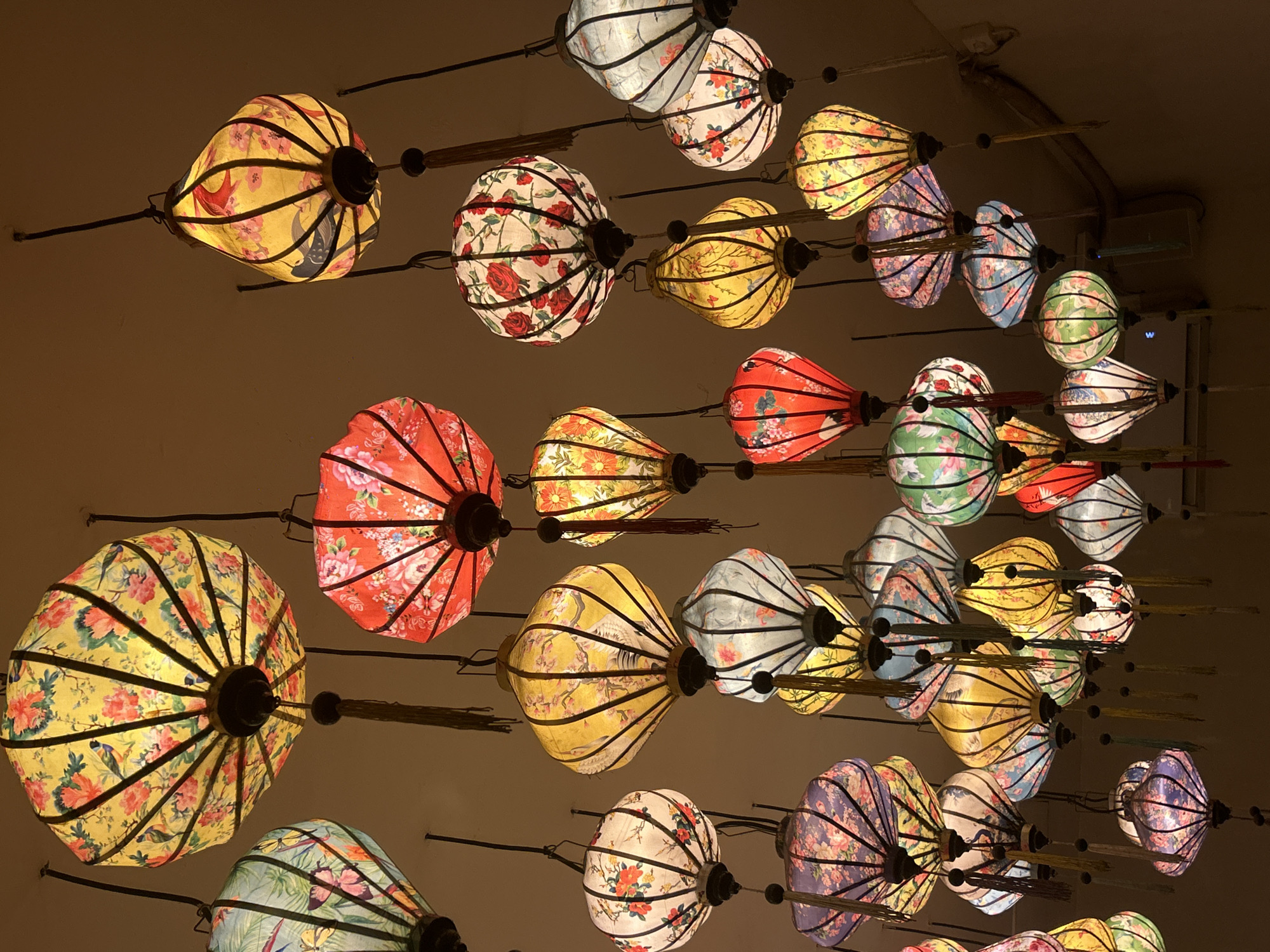 The image depicts a collection of brightly lit, decorative floral lanterns hanging from a ceiling. The lanterns are spherical and shaped like flower buds, with a variety of colors including yellow, red, blue, green, and white. The location is likely in North Kuta, Indonesia based on the context.