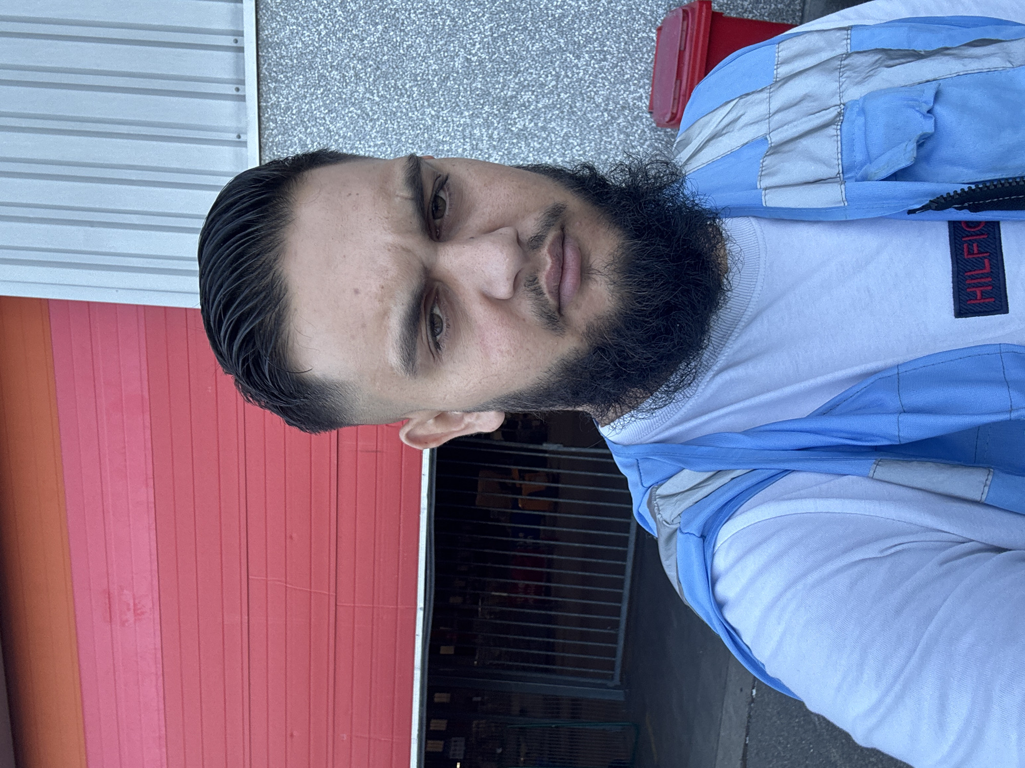 A man with dark hair styled in a fade and a full beard is taking a selfie outdoors. He is wearing a white t-shirt with a navy Hilfiger logo and a light blue and gray reflective vest. Behind him, a large red industrial roll-up door is partially open, revealing a dark interior with metal gates. To the right, a speckled gray building wall is visible, along with a red trash bin. The man's expression is neutral. The environment suggests an industrial or commercial area, potentially a warehouse or loading dock. The city of Boom, Belgium, is known for its industrial heritage and is often associated with port activities and large logistical centers, which aligns with the visual cues in the image.
