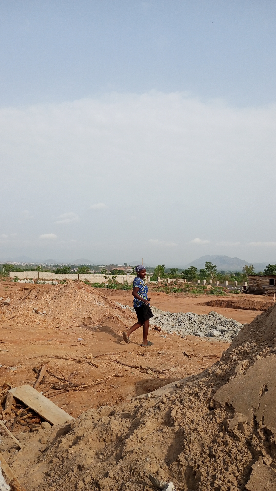 A medium-shot captures a woman walking across a dirt and construction-filled landscape on the outskirts of Zango-ndaji, Nigeria. The woman wears a patterned blue shirt, dark shorts, and sandals, with a head wrap. She walks from left to right. The ground is predominantly red dirt, with piles of dirt and gravel interspersed. A stack of grey rubble lies near the center of the frame. Some scrap wood and other construction debris are scattered around. Concrete wall segments stand in the background, beyond the building site, indicating some degree of development. Further in the distance, the cityscape of Zango-ndaji is visible, with hills rising beyond it. The sky is overcast and pale blue.