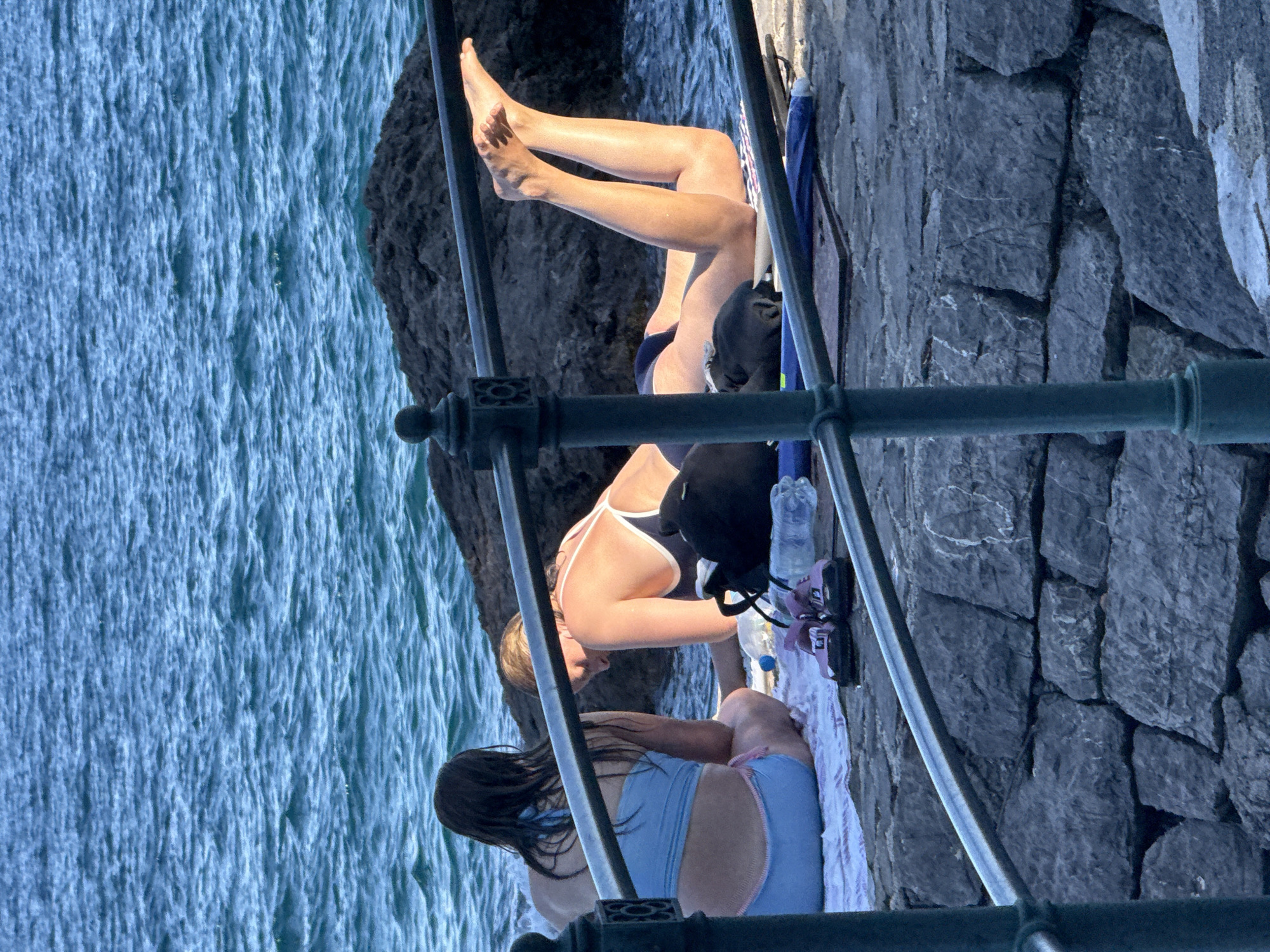 Two individuals are sunbathing on a stone quay in Opatija, Croatia. The person on the left is seated facing away from the camera, wearing a light blue bikini. The person on the right is lying down, facing towards the sea, wearing a dark swimsuit with white straps. Their legs are raised and their feet are visible. A black bag and a pair of purple sandals are placed on a towel on the ground. The water is choppy and blue. A dark rock formation is visible in the background, and a metal railing runs horizontally across the foreground. The quay is constructed of large, grey stones.