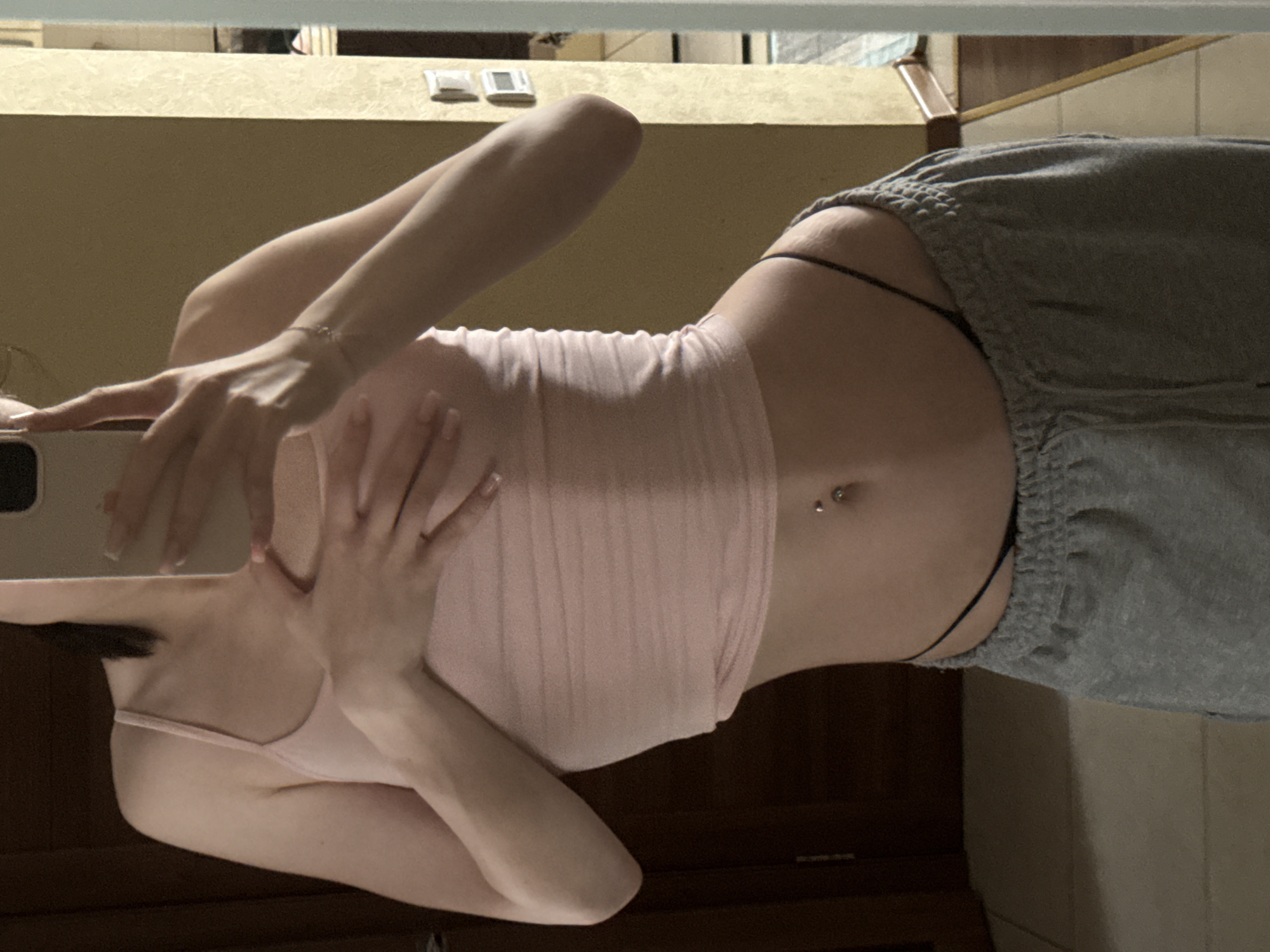 In Krasnogorsk, Russia, a young woman is captured in a selfie taken in a dimly lit room, likely her home. She stands in front of a mirror, her reflection filling the frame. She is wearing a light pink tank top that exposes her midriff, and grey sweatpants. A black thong peeks out from the waistband of her sweatpants. Her left hand is placed over her chest, while her right hand holds her phone, which is partially obscuring her face. A navel piercing is visible on her stomach. The background shows a dark wooden door on the left and a textured wall with light switches on the right. The lighting suggests it is either early morning or evening.