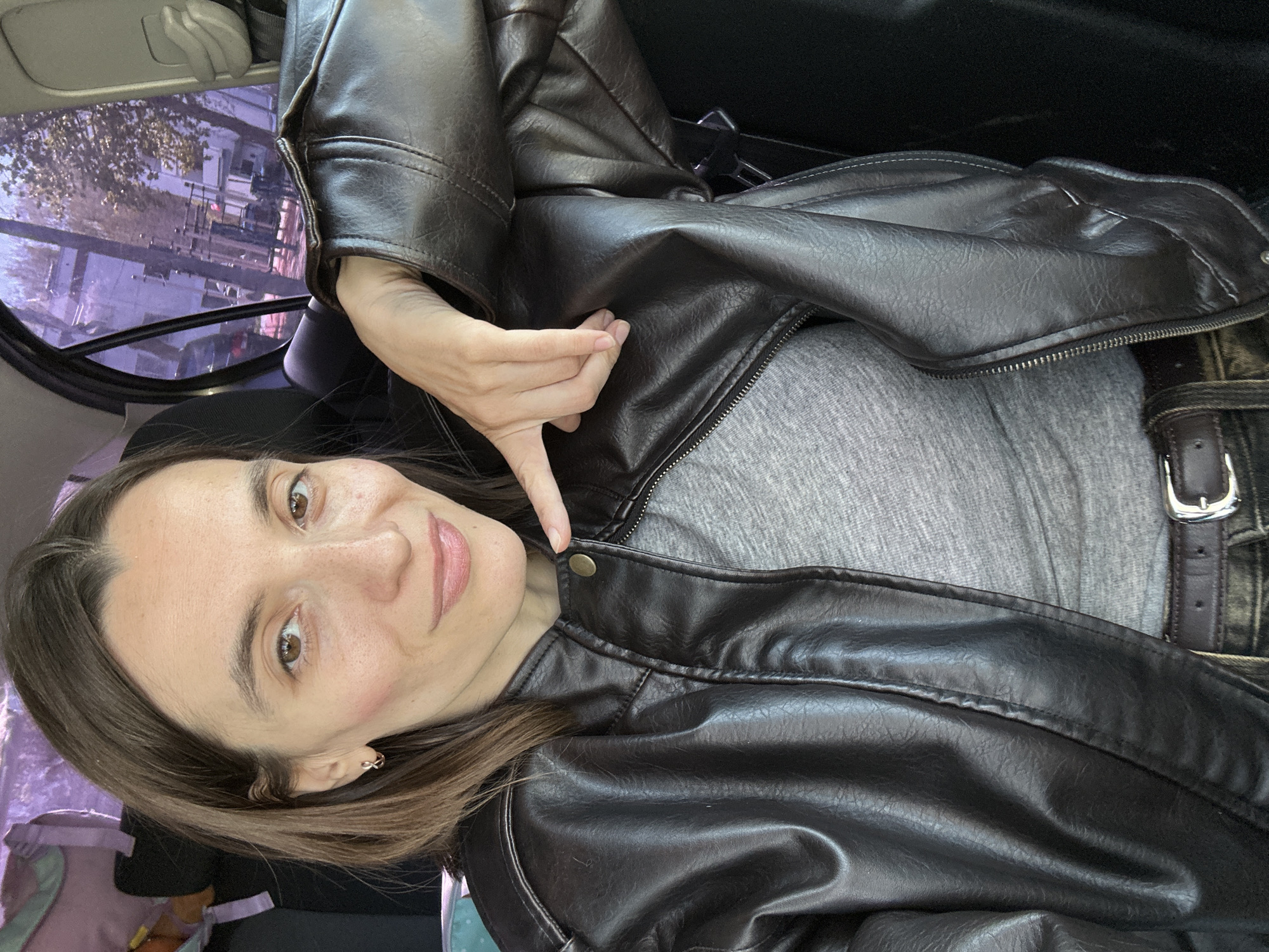 A woman with long brown hair is in a car, wearing a grey shirt and a black leather jacket. She is holding up her index finger, pointing toward her throat. Outside the car window, a blurred street scene is visible, with buildings and trees. The exact location in Moscow, Russia, is not discernible from the provided image.