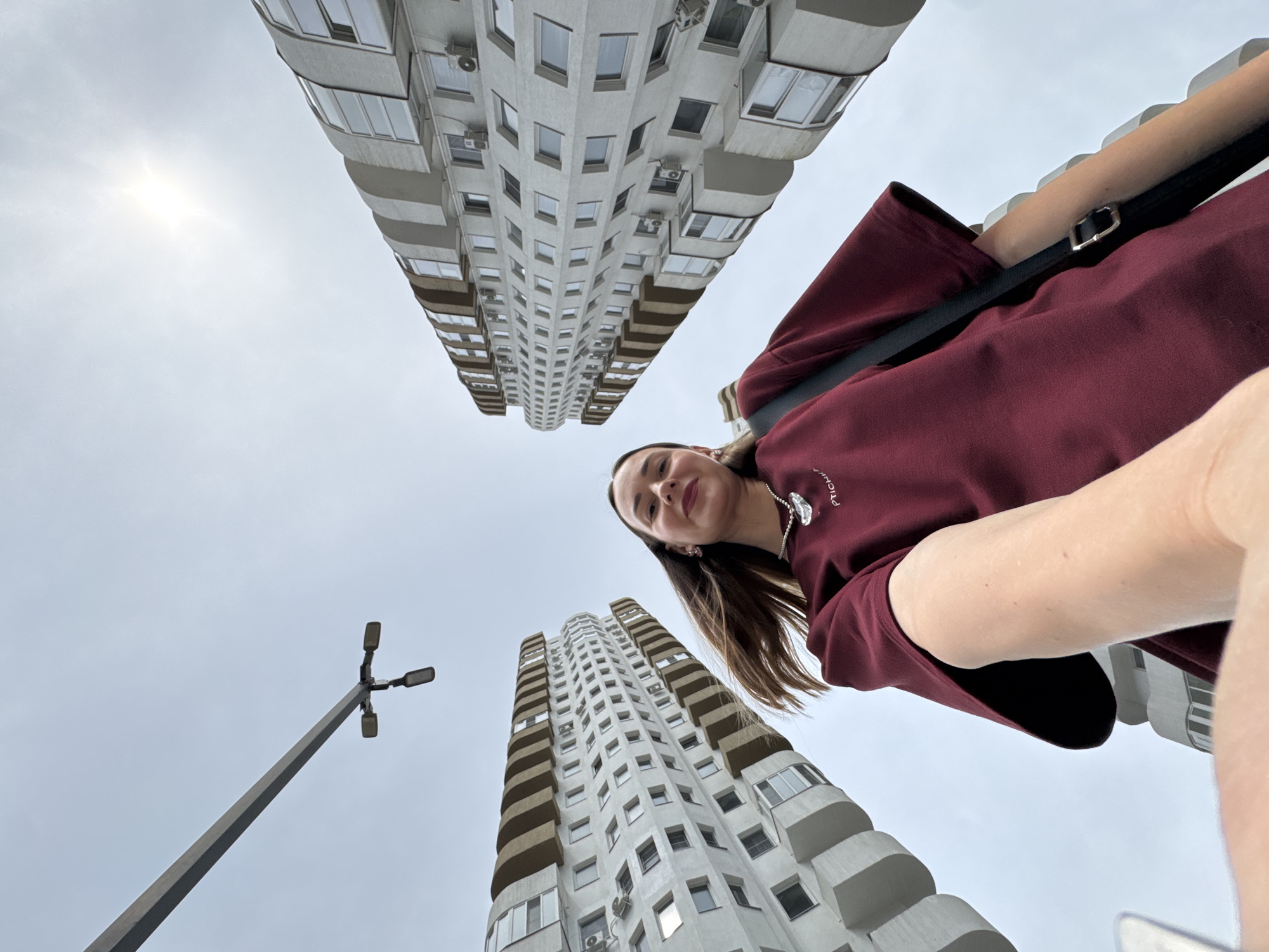 A low-angle, wide shot captures a young woman taking a selfie outdoors in front of tall, modern residential buildings. The woman, with long brown hair, is wearing a dark red t-shirt and a silver necklace. Her right arm is extended, holding the camera. Behind her, two towering buildings with white facades and curved balconies dominate the background. A street lamp is visible in the upper left quadrant of the image, and the sun is peeking through a cloudy sky. The image appears to have been taken in Sochi, Russia, given the architectural style of the buildings.
