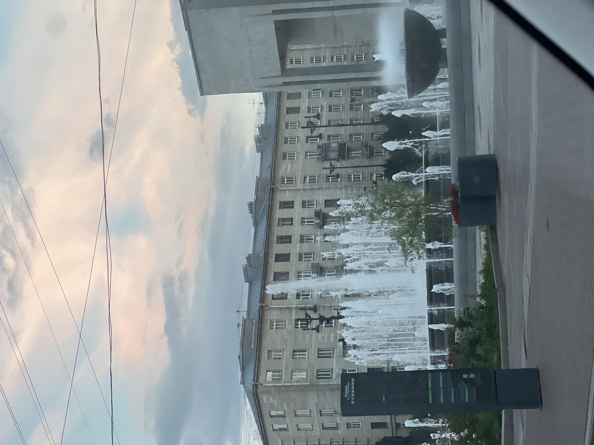 The image shows a large fountain with multiple jets of water in front of a multi-story apartment building with a gray facade. The building has numerous windows and balconies. To the right of the fountain, there is a large, modern, gray structure with an archway. The sky is partly cloudy with soft pink and gray hues, suggesting it may be dawn or dusk. Several overhead wires are visible against the sky. In the foreground, there is a dark, rectangular information kiosk. The area around the fountain appears to be a public space with a paved ground. This scene is likely located in Saint Petersburg, Russia, given the architectural style and the presence of overhead wires, which are common in older urban areas of the city.