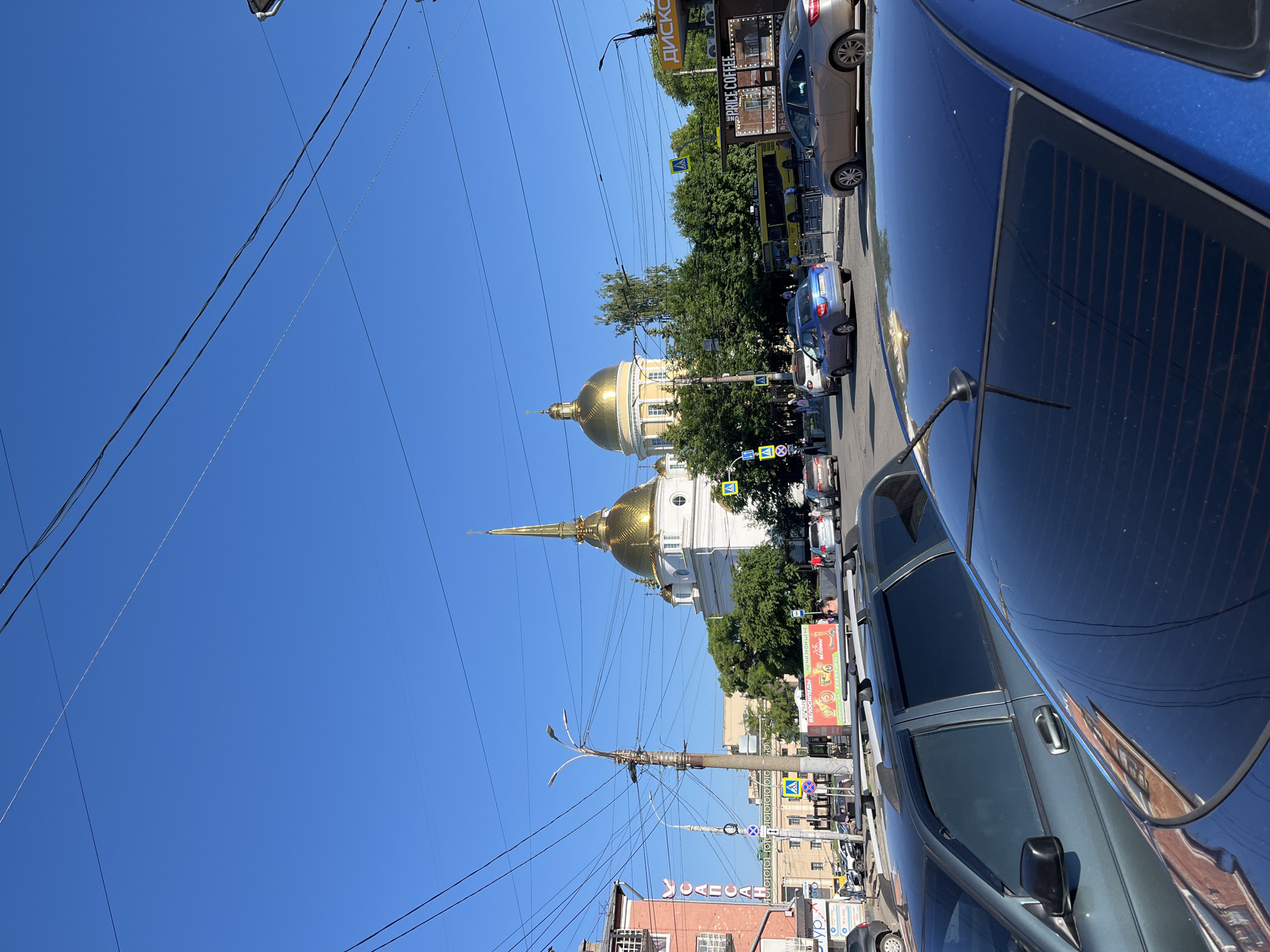 The image captures a sunny day in Izhevsk, Russia, likely a city street with moderate traffic and pedestrian activity. Dominating the scene is the Kazan Cathedral, a prominent landmark with its striking golden domes, visible behind a row of trees. The cathedral's white facade and ornate architecture stand out against the clear blue sky.
In the foreground, several cars are parked, including a dark blue hatchback and a dark gray station wagon. The street is busy with traffic, with cars and a yellow bus moving along. Pedestrians are scattered throughout the scene, some crossing the street at designated crosswalks, marked with yellow pedestrian signs.
On the right side of the image, a sign with the text "ДИСКО" and "TERRACE COFFEE" is visible, suggesting commercial establishments in the vicinity. A bus stop shelter is also present, with people waiting for public transport. The street is lined with buildings, some of which appear to be older structures with a distinct architectural style. Overhead, a complex network of electrical wires crisscrosses the sky, a common feature of urban landscapes.
The overall impression is a snapshot of everyday life in a Russian city, with a blend of historical architecture, modern infrastructure, and bustling urban activity. The sunlight casts sharp shadows, indicating a bright and clear day.