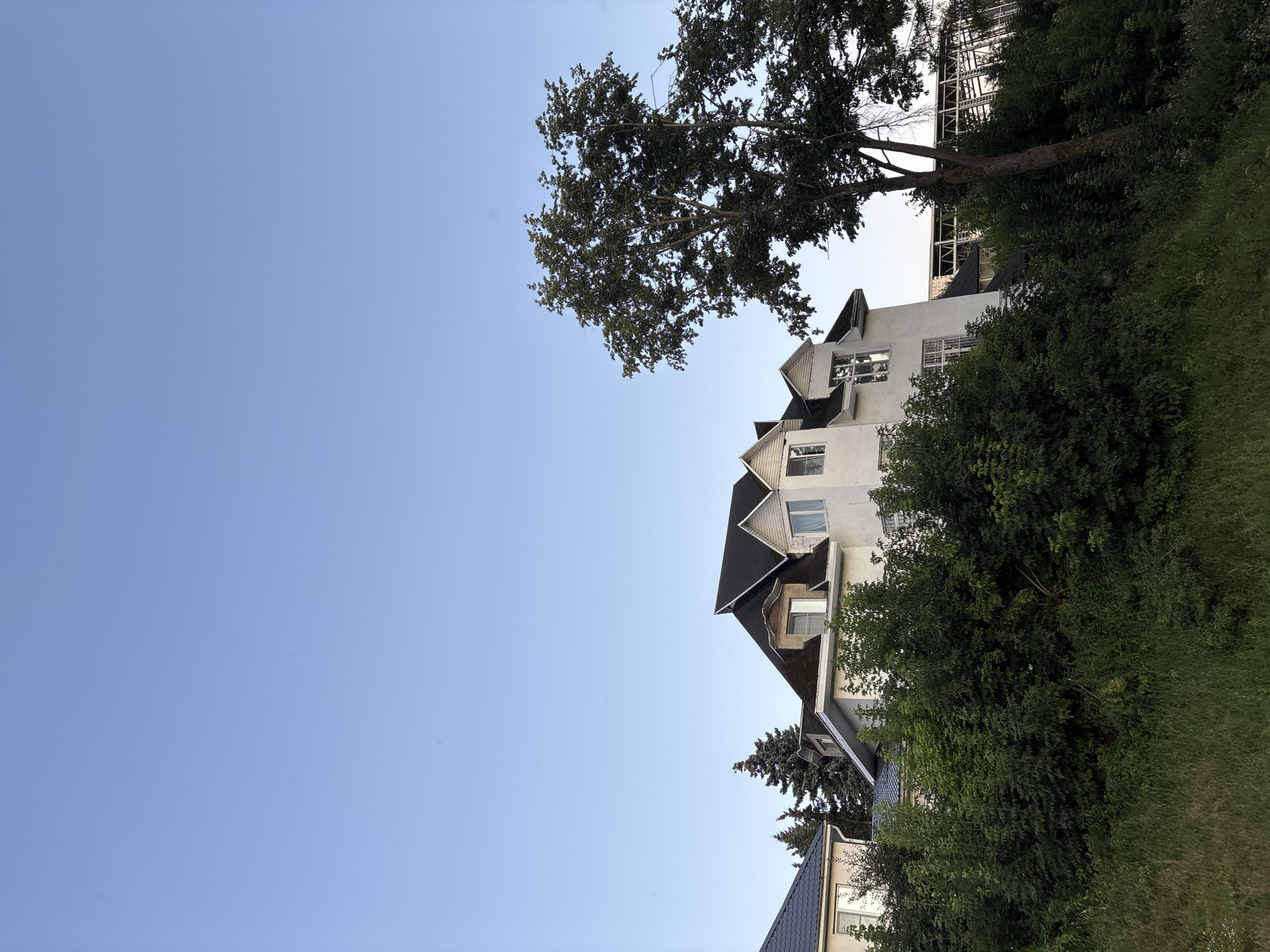 A bright, clear sky dominates the upper portion of the image. Below, a multi-story white house with a dark roof and several dormer windows is visible. The house is partially obscured by lush green trees and bushes in the foreground. To the left, the roof of another house with a dark, tiled roof can be seen. A large tree with dark green foliage stands to the right of the white house, its branches extending into the sky. In the background, behind the trees and the white house, a partially constructed or deconstructed building with a metal frame structure is visible. The overall impression is of a residential area with significant vegetation, set against a backdrop of construction or renovation. The specific location within the city of Klin, Russia is not identifiable from the image itself.