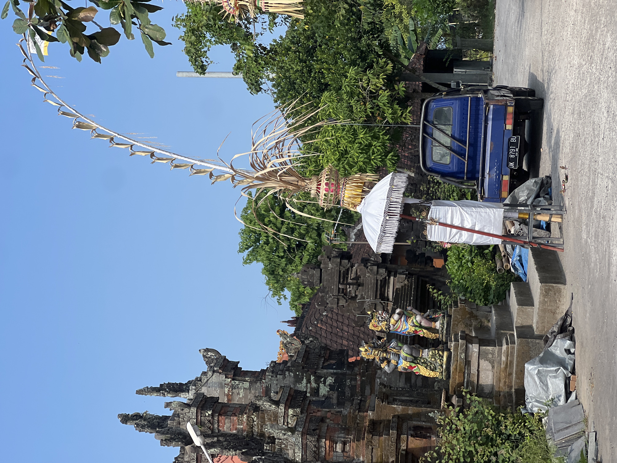 The image depicts an outdoor scene in Kediri, Indonesia, featuring traditional Balinese temple architecture and elements associated with local culture and ceremonies. In the foreground, a blue Suzuki pickup truck is parked on a paved road. To the left of the truck, a white umbrella with fringe detail stands upright, adorned with draped white fabric and decorative elements. Further left, two stone statues, likely representing temple guardians or deities, are positioned in front of a stone staircase leading up to a traditional brick temple structure with intricate carvings and a tiled roof. The temple is partially overgrown with vegetation. Above and behind the temple, lush green trees and a clear blue sky dominate the background. A tall, curved bamboo structure, possibly part of a festive decoration or offering, arches over the scene, adorned with dried leaves and woven baskets. Other decorative elements, such as hanging ornaments, are also visible among the trees. The overall impression is one of a cultural or religious site prepared for an event.