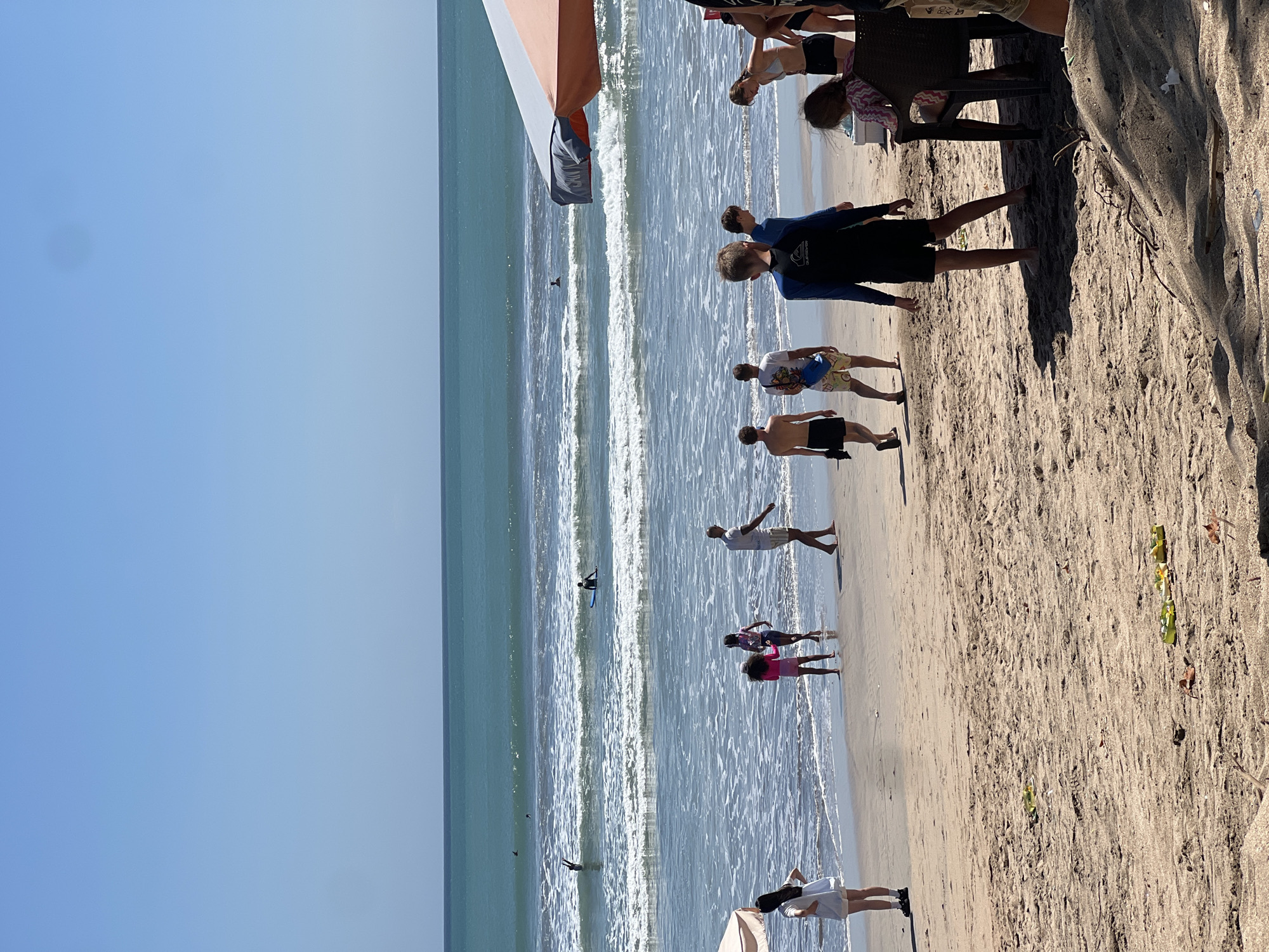 The image depicts a sunny day at Kuta Beach, Bali, Indonesia, with a clear blue sky and moderate waves. Several people are present on the sandy shore and in the water. In the foreground, on the sand, a group of individuals are walking and standing. Two children in pink and purple swimsuits are near the water's edge, while an adult male in white shorts walks away from the ocean. A woman in a white dress is also visible on the left. Further back, a group of young people, including boys wearing rash guards and swim trunks, are seen walking. In the ocean, several surfers are visible. One individual is on a surfboard in the middle ground, and another appears to be body surfing. Further out, two more figures are in the water. The beach is populated with a few chairs and umbrellas, suggesting a typical beach scene. The overall atmosphere is one of leisure and recreation.