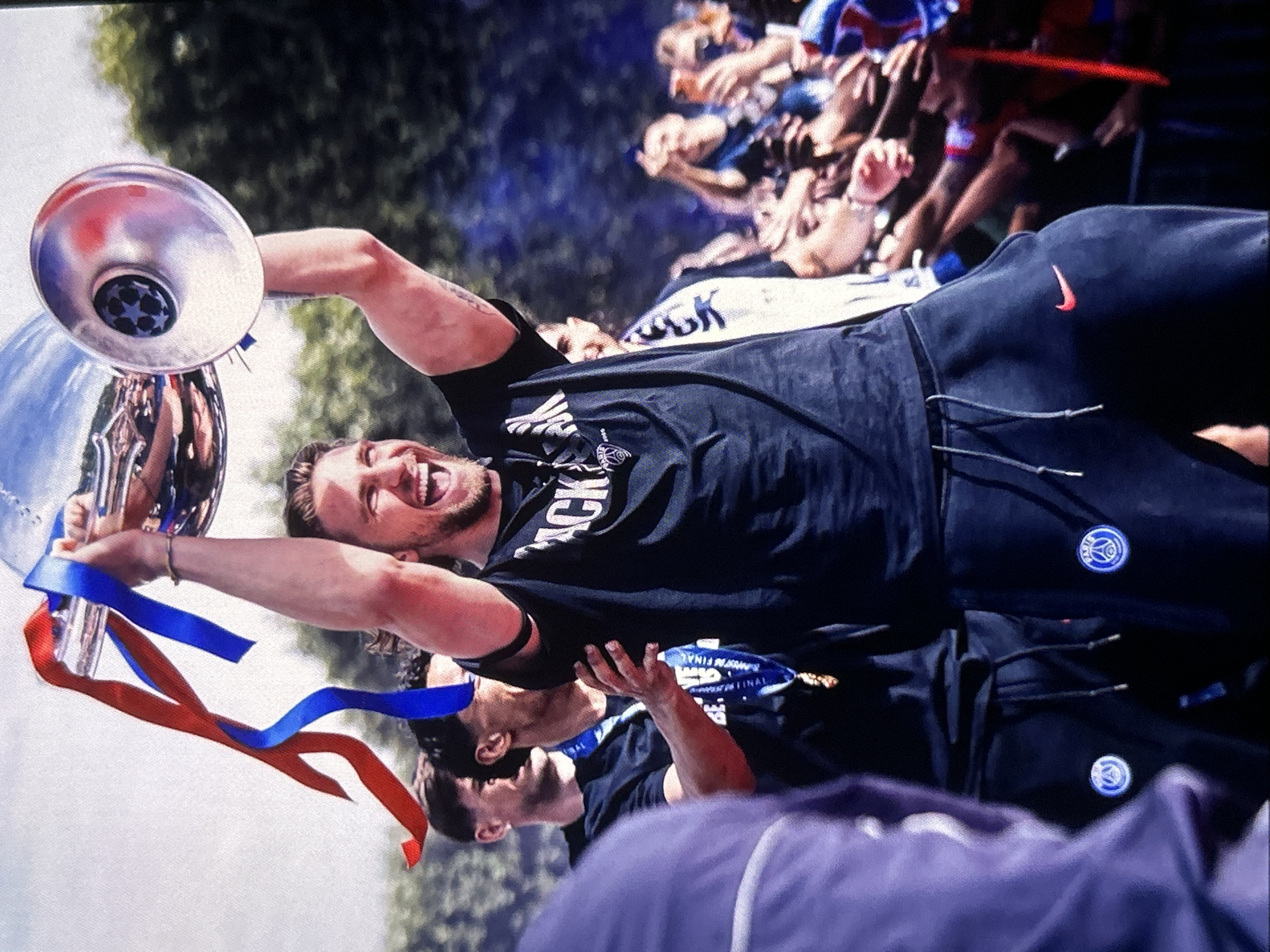 The image captures a vibrant moment of celebration, likely a victory parade, with a main focus on a joyful man holding a trophy. This event is contextualized as taking place in Bania, Bulgaria.
The central figure is a man with a beard, wearing a black t-shirt with "BLACK" partially visible across the chest, and black tracksuit bottoms. He is mid-laughter or cheer, with his mouth wide open, and his arms raised high as he hoists a large, gleaming silver trophy. Red and blue ribbons are tied to the trophy, which also features a dark blue circular emblem with white stars, consistent with the UEFA Champions League logo.
The man's tracksuit bottoms have a red Nike swoosh logo on the right thigh and a circular blue and white club crest on the left thigh. Below him, another man is partially visible, also in black attire with a similar blue and white crest on his pants. He wears a blue medal or ribbon around his neck, which clearly displays the words "CHAMPION FINAL" and "UEFA".
In the blurred background, a crowd of enthusiastic fans can be seen with hands raised, participating in the celebration. Green foliage from trees indicates an outdoor setting, suggesting a bright and sunny daytime event. The overall emotion conveyed is one of immense joy, triumph, and shared excitement between the players and their supporters. The image itself is rotated counter-clockwise.