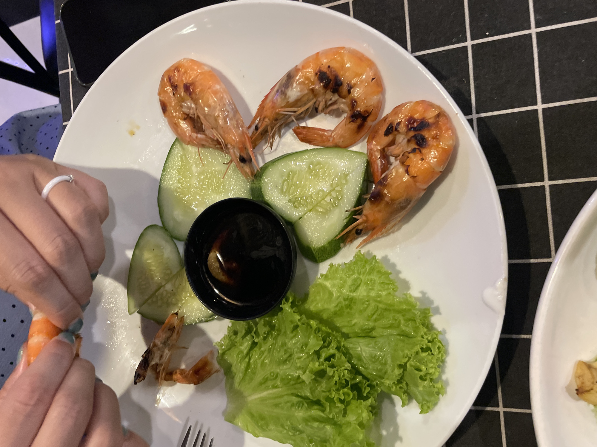 A white plate, resting on a black and white checkered surface, contains grilled shrimp, cucumber slices, lettuce, and a small black bowl filled with sauce. A person with a ring on their finger is peeling a shrimp with their hands. The scene is likely located in or near Nha Trang, Vietnam, as indicated by the prompt's location specification. The image depicts someone preparing to eat a meal featuring seafood and fresh vegetables, with a dipping sauce. A fork is visible at the bottom of the plate. Another partially visible plate is visible to the right.
