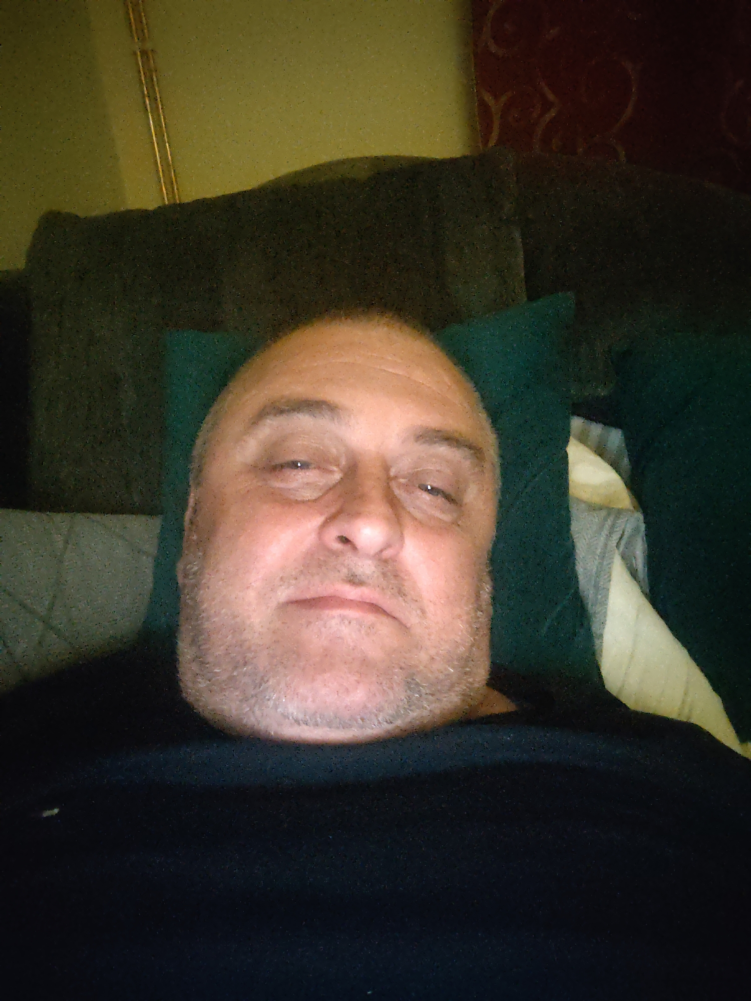 This image depicts a single adult male, presumed to be located in Huntingdonshire, United Kingdom, captured from a low-angle perspective while he is lying down. The Caucasian subject, appearing middle-aged with a receding hairline and short facial stubble, wears a dark, possibly black, crew-neck shirt. His head rests on a dark teal or green pillow, and he looks directly into the camera with a relaxed or tired expression, exhibiting partial lid closure on his right eye. The immediate background consists of dark green upholstered furniture or bedding. Further background elements include a light green wall with a vertical metallic strip on the left and a reddish-brown patterned wall on the right. The scene is dimly lit, resulting in noticeable image noise.