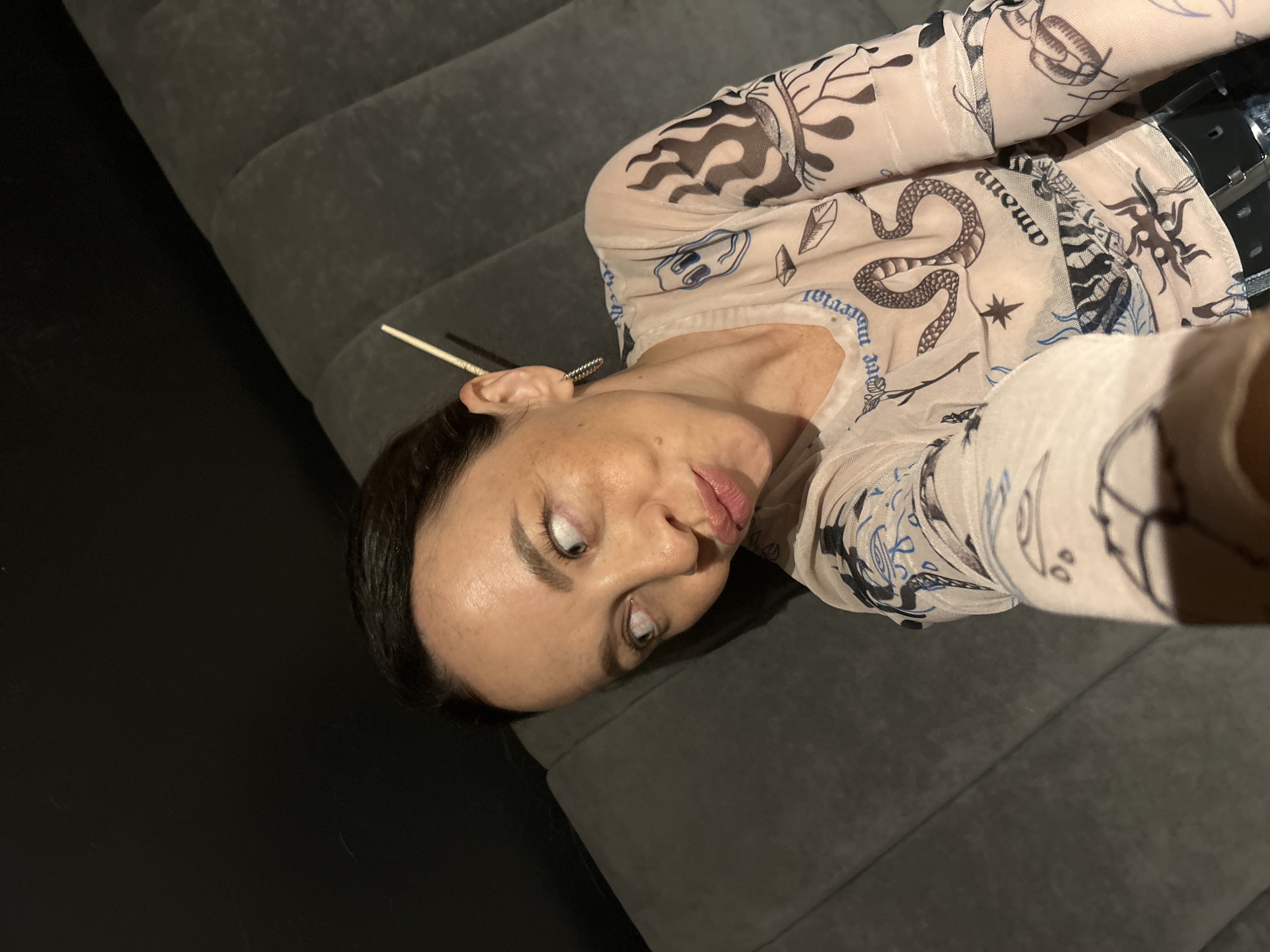 A woman with dark hair pulled back is looking to her left. She is wearing a sheer, long-sleeved shirt with a pattern of snakes, flames, and faces. A chopstick is sticking out from behind her ear. She appears to be sitting on a dark gray, tufted couch. The background is dark and out of focus. There is no indication of the city of Chaikovsky, Russia, in the image.