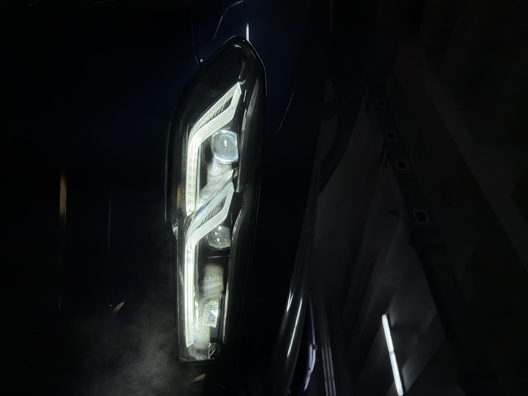 The image shows a close-up of a car headlight at night. The headlight is illuminated and features a modern LED design with a complex arrangement of light elements. The car appears to be dark or black. The background is indistinct, with some blurry shapes and light sources. The photo may have been taken indoors or in a dark outdoor setting. There is no information in the image to suggest that it was taken in Krasnoyarsk, Russia. There are no visible people, subjects, actions, or interactions in the image.
