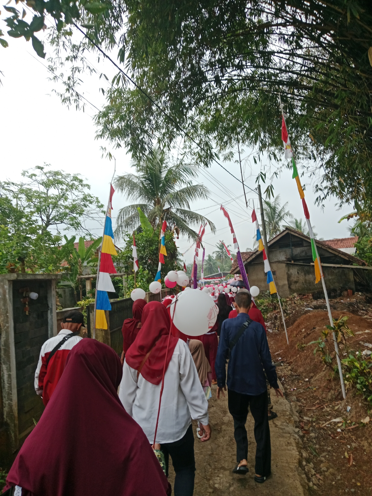 In Bojongmengger, Indonesia, a procession moves along a narrow paved path lined with trees, flags, and houses. Several individuals are visible, most wearing traditional Islamic attire, including headscarves in red and maroon. Some are carrying large white balloons. The path is flanked by vegetation and a building with a weathered brown roof. Red and white flags, along with flags of other colors like blue, yellow, and green, are displayed on poles, suggesting a festive or celebratory occasion. The sky is overcast, contributing to a subdued lighting.