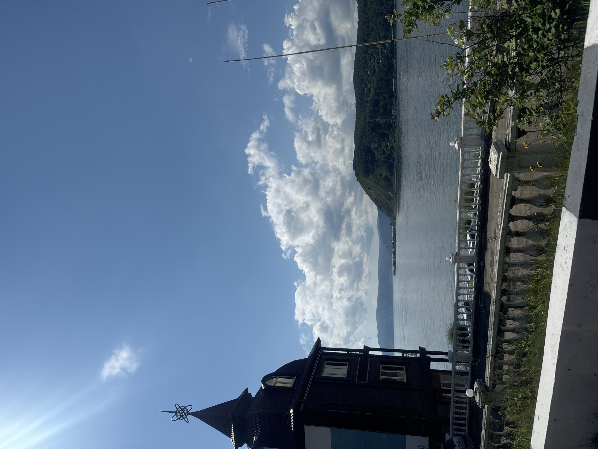 The image shows a view of Lake Baikal from the shore in Listvyanka, Russia. In the foreground, there is a dark, multi-story building with a distinctive pointed roof topped with a decorative element resembling an astrolabe. The building has several windows and balconies. In front of the building, a white stone balustrade runs along the edge of the property, with the lake visible beyond. The lake is calm and reflects the blue sky, which is dotted with fluffy white clouds. On the far side of the lake, a lush green, tree-covered hill rises steeply, with a few scattered buildings visible on its slopes. The scene is bathed in bright sunlight, with a noticeable lens flare in the upper left corner of the image. The overall impression is of a scenic, peaceful waterside location.