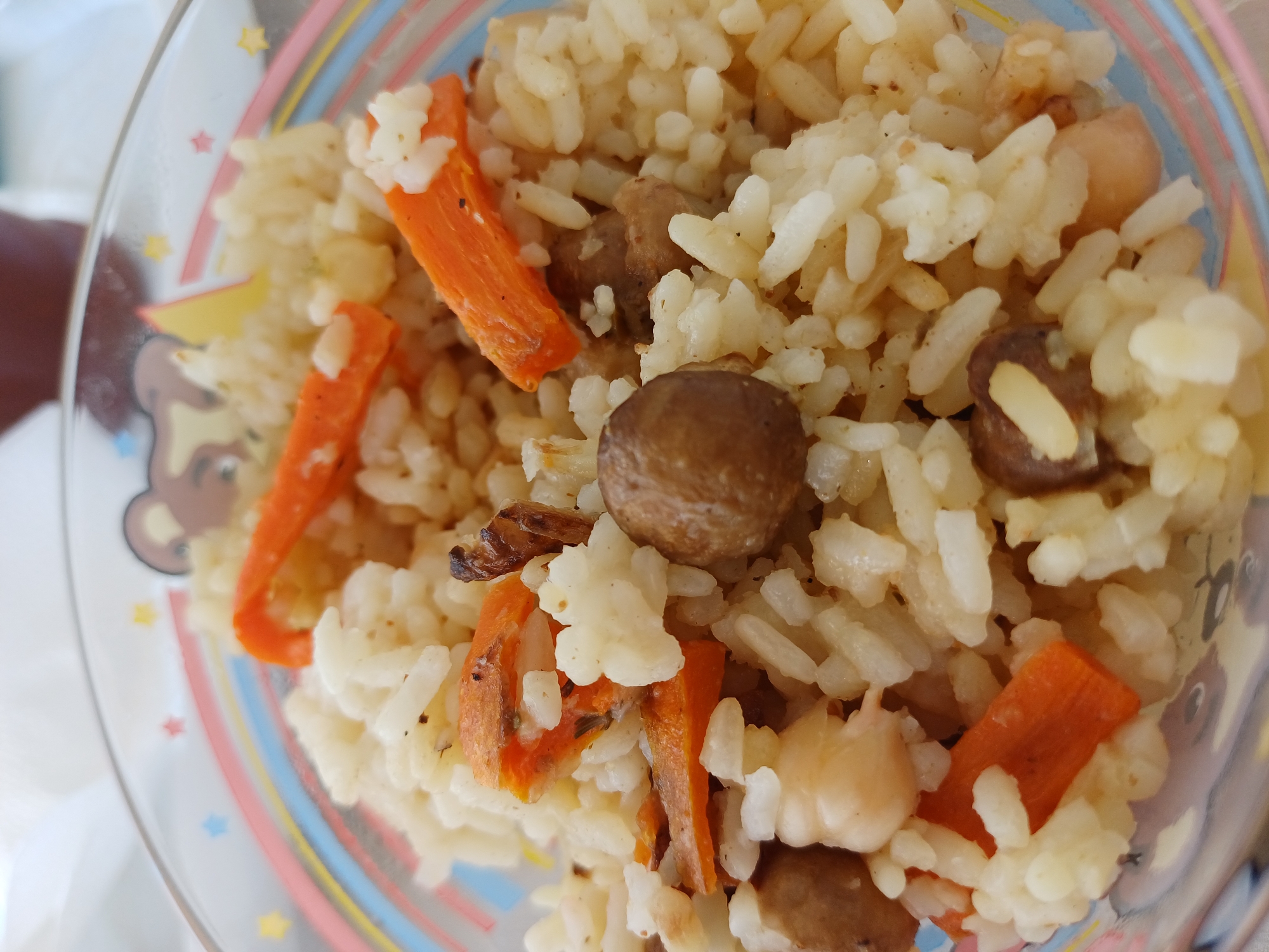 This is a close-up, overhead shot of a bowl of pilaf, a rice dish, served in a clear glass bowl. The pilaf contains white rice, cut carrots, and what appear to be chickpeas and possibly small pieces of meat or mushrooms. The bowl itself has a decorative rim with a pattern of colorful stripes and cartoonish images of stars and a bear. The lighting suggests it is daytime, likely indoors, and the overall impression is of a home-cooked meal. No people are visible, and the focus is solely on the food. There is no visible text in the image. The location context provided is Sochi, Russia, but this cannot be visually confirmed from the image itself. The image depicts a food item prepared for consumption.