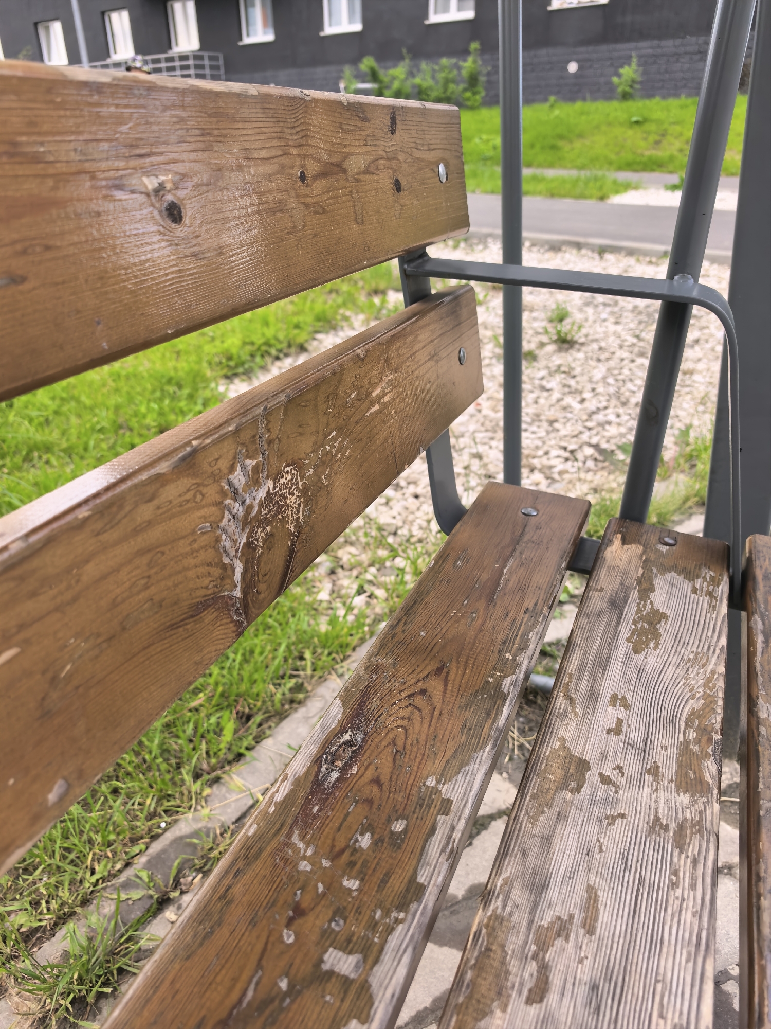 This media file is a close-up shot of a weathered wooden bench, likely part of a swing set or park furniture. The bench is made of dark brown wooden planks with visible grain patterns and signs of wear, including peeling paint or varnish and some darker, possibly wet, patches. The wood is fastened with silver screws.
The bench is positioned outdoors, with a background of green grass, a paved walkway, and a modern, dark-colored building with white windows. The scene suggests an outdoor setting during the daytime, with the lighting indicating it is likely overcast or shaded, as there are no strong shadows. The overall impression is one of an outdoor public space that has seen some use. There is no visible text in the image.