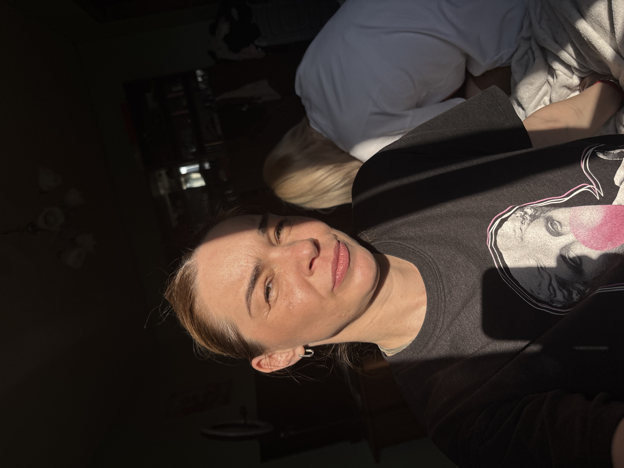In an indoor setting, possibly a bedroom, in Vsevolozhsk, Russia, a woman with light brown hair pulled back and wearing a dark t-shirt with a graphic print is captured in a selfie. The low-angle shot shows her from the chest up, with sunlight casting a shadow diagonally across her face and shirt. Her expression is neutral, with her eyes looking towards the camera. She is wearing a small hoop earring in her left ear. To her right and slightly behind her, another person with blonde hair, wearing a white t-shirt, is partially visible, facing away from the camera and appearing to be leaning over. The background is dimly lit, with hints of furniture and shelves suggesting a domestic environment. The overall lighting creates a dramatic contrast between light and shadow.
