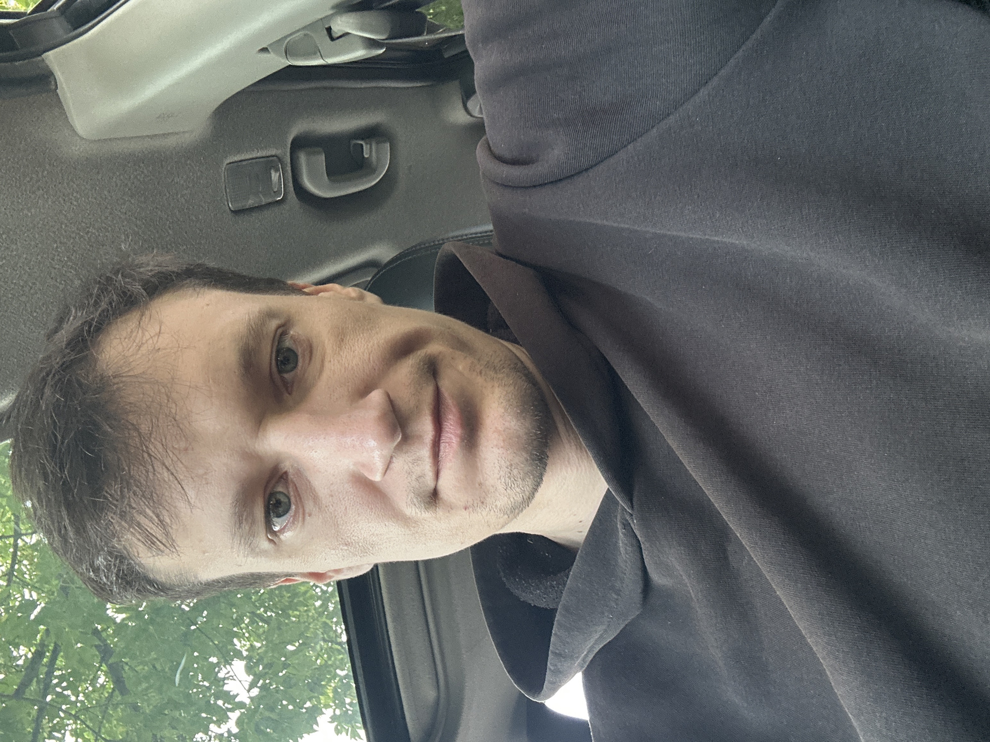 A medium shot selfie shows a man with light skin, brown hair, and blue eyes looking directly at the camera. He is wearing a dark gray hooded sweatshirt. He is inside a vehicle, with the interior visible behind him, including a sunroof and parts of the car's ceiling and door frame. The background also shows a glimpse of green foliage, suggesting an outdoor environment. The man's expression is neutral.