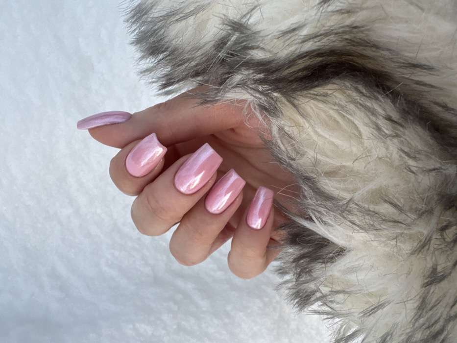 A close-up image depicts a human left hand with five fingers visible. The hand, positioned with fingers slightly fanned, features elongated, coffin-shaped fingernails painted with a pearlescent light pink polish. The hand rests on a textured surface of faux fur, predominantly off-white with dark grey and black strands. The background is a soft, white, indistinct textured material. Location: Vniissok, Russia.