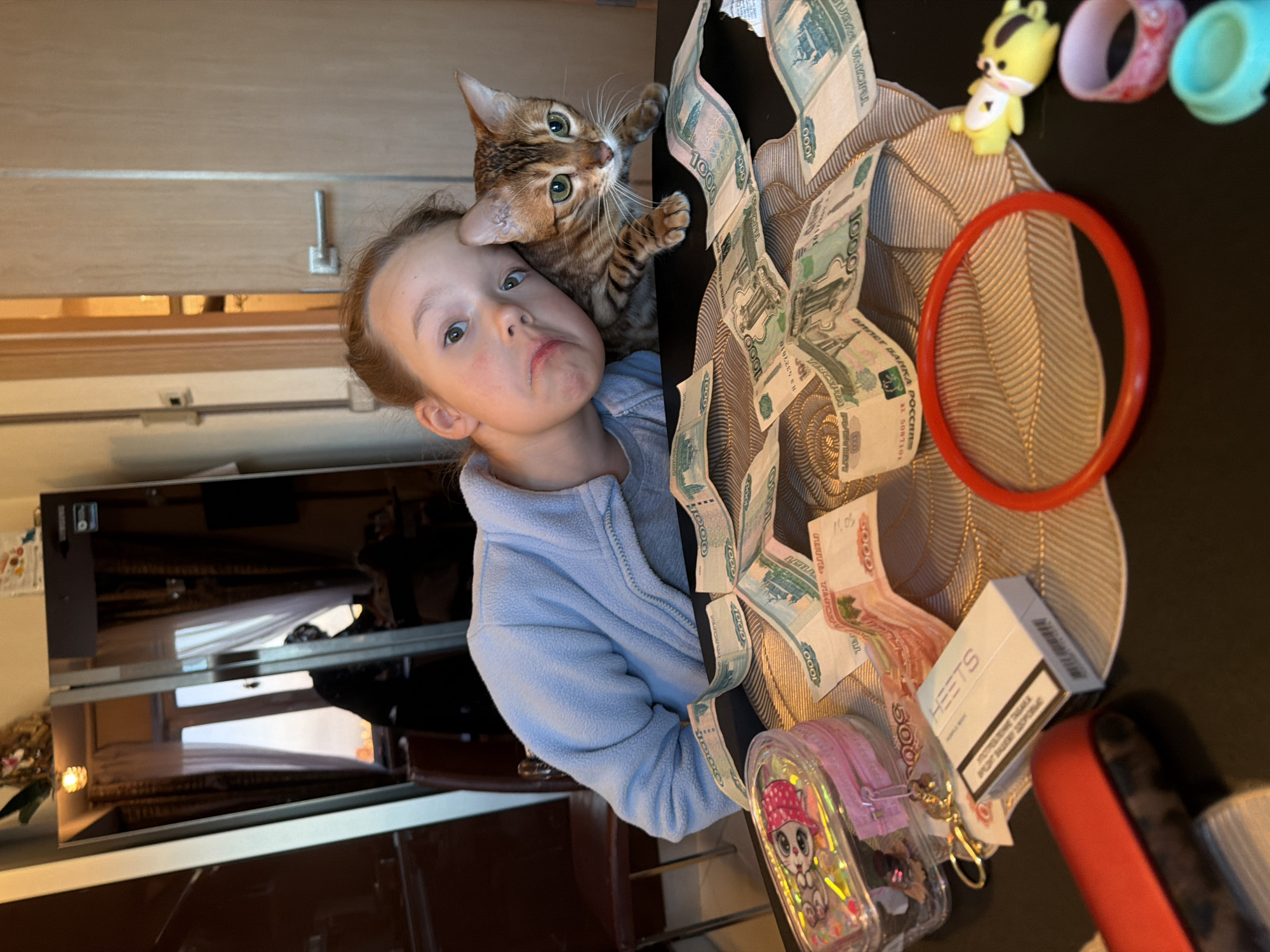 The image depicts an indoor scene in a residential environment, identified as Saint Petersburg, Russia. A young girl, approximately 5-10 years old, with light brown hair, wears a light blue fleece jacket and leans her head to her left, looking directly at the camera with a slight pout. Adjacent to her, on a dark table, is a Bengal cat with spotted fur and green eyes, also looking towards the camera.
The table features a light-colored placemat with a textured leaf pattern, upon which numerous Russian Ruble banknotes are scattered; specifically, 1000 Ruble (green/blue) and 5000 Ruble (red/pink) denominations are visible. To the left of the currency, a clear plastic container with a pink zipper and a cartoon cat design holds small items, with a set of keys attached. A white box of "HEETS" brand heatsticks, bearing Russian text that includes a health warning, rests near the money. A solid red circular bangle is present on the right side of the table, accompanied by a yellow animal-shaped toy and two small, colorful rings (one pink, one light blue). A red electronic device or case is partially visible on the far left.
The background on the left is dominated by a tall, reflective dark surface, consistent with a refrigerator or mirrored cabinet. This surface reflects an interior window with both sheer white and dark patterned curtains, through which an outdoor sky is visible. A dark brown chair with a metal frame is partially visible behind the girl. On the right side of the background, a light-colored wooden door with a silver handle is present. An ornamental light fixture or object is positioned atop the reflective surface in the upper left.