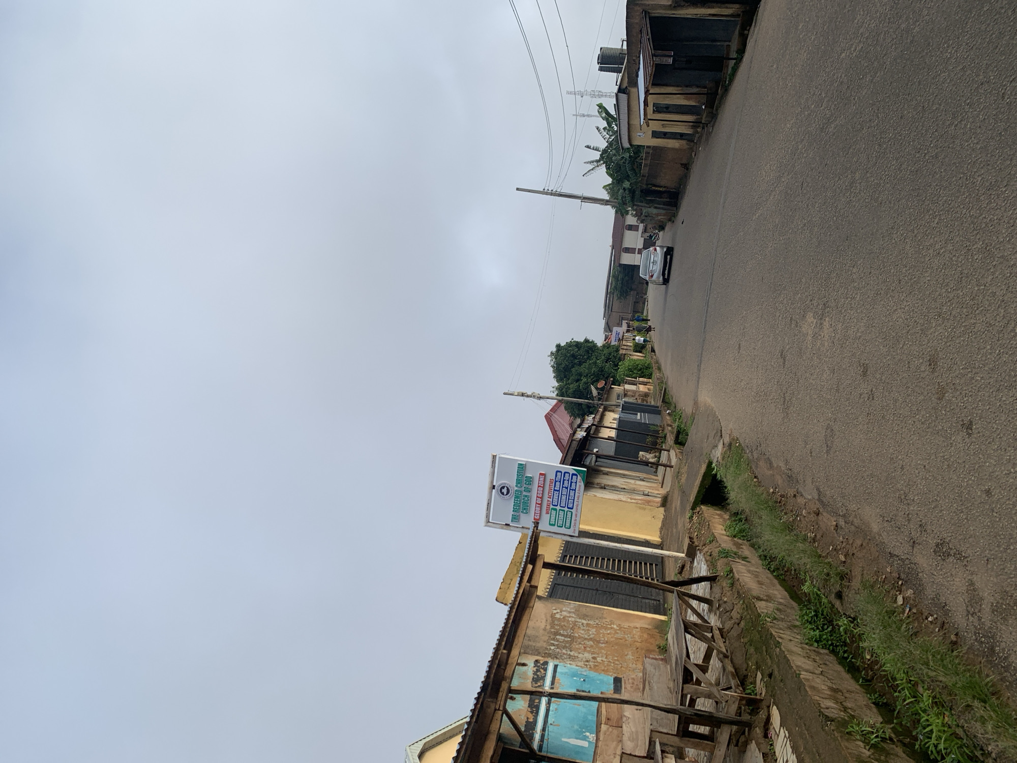 This is a photograph taken in Jos, Nigeria, showing a street scene on an overcast day. The image is oriented vertically, with the road running diagonally from the bottom right to the top left. On the left side of the road, a row of buildings and structures are visible. One building has a prominent sign for "THE REDEEMED CHRISTIAN CHURCH OF GOD GLORY OF GOD ZONE," detailing its weekly activities and service times. The structures appear to be a mix of older, somewhat dilapidated buildings and more modern constructions. Some of the buildings have corrugated metal roofs and weathered walls.
A paved road occupies the right side of the frame, with a narrow, overgrown drainage ditch running alongside it. A single white car is seen driving on the road in the distance, and a few indistinct figures are visible walking near the buildings on the left. Overhead, a network of power lines stretches across the sky, supported by utility poles. The overall impression is of a typical urban or semi-urban environment in Nigeria, with a cloudy, possibly humid, atmosphere. The time of day is not explicitly indicated, but the overcast sky suggests it could be morning or afternoon.
