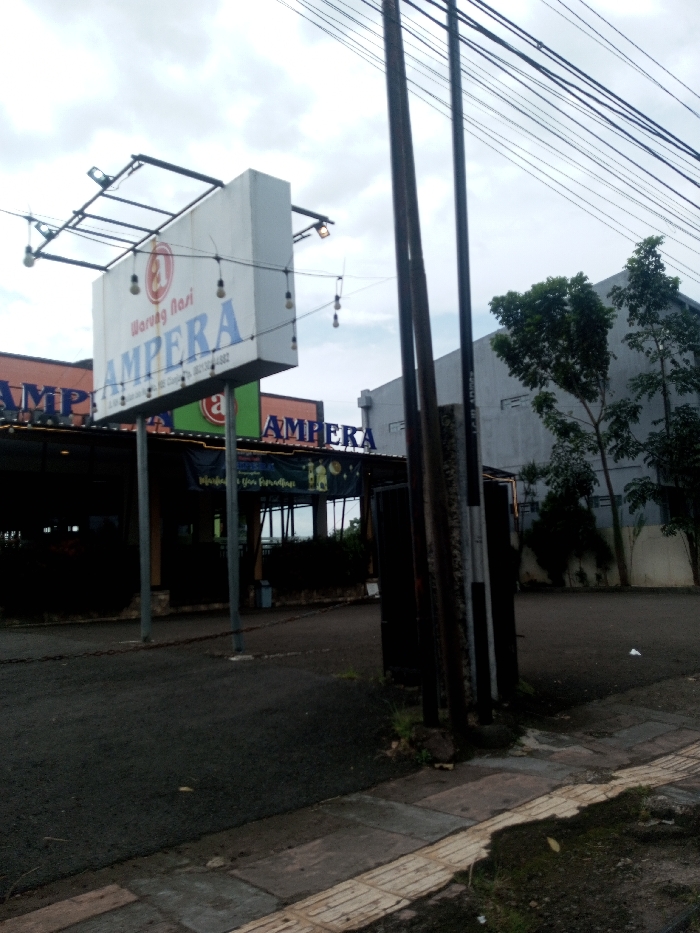 The image depicts an outdoor commercial scene in the city of Chhenangri_Rishor, Bhutan, under an overcast sky. A prominent white rectangular sign, supported by a metal frame, dominates the mid-left, displaying "Warung Nasi" above a circular logo, and "AMPERA" in large blue lettering below. Smaller text on the sign includes what appears to be a street address and phone number "0821-1254-6882". String lights with exposed bulbs are draped around the sign and its frame. Directly beneath this sign, an establishment with an orange facade and another "AMPERA" sign is partially visible, appearing to be an open-air or semi-enclosed dining area. A blue banner, possibly related to "Ramadhan," is strung across the front of this structure.
On the right side of the frame, three dark utility poles rise vertically, supporting numerous black electrical or communication wires that traverse the upper portion of the scene. Behind these poles, a large, light grey building with a flat roof is visible, flanked by several trees with green foliage. The ground in the mid-ground consists of a dark asphalt surface, likely a parking area or access road. In the immediate foreground on the right, a section of a concrete paver sidewalk is visible, featuring a yellowish tactile paving strip along its edge. Sparse vegetation and dirt are present along the ground's edges. No individuals or discernible activities are observed.
