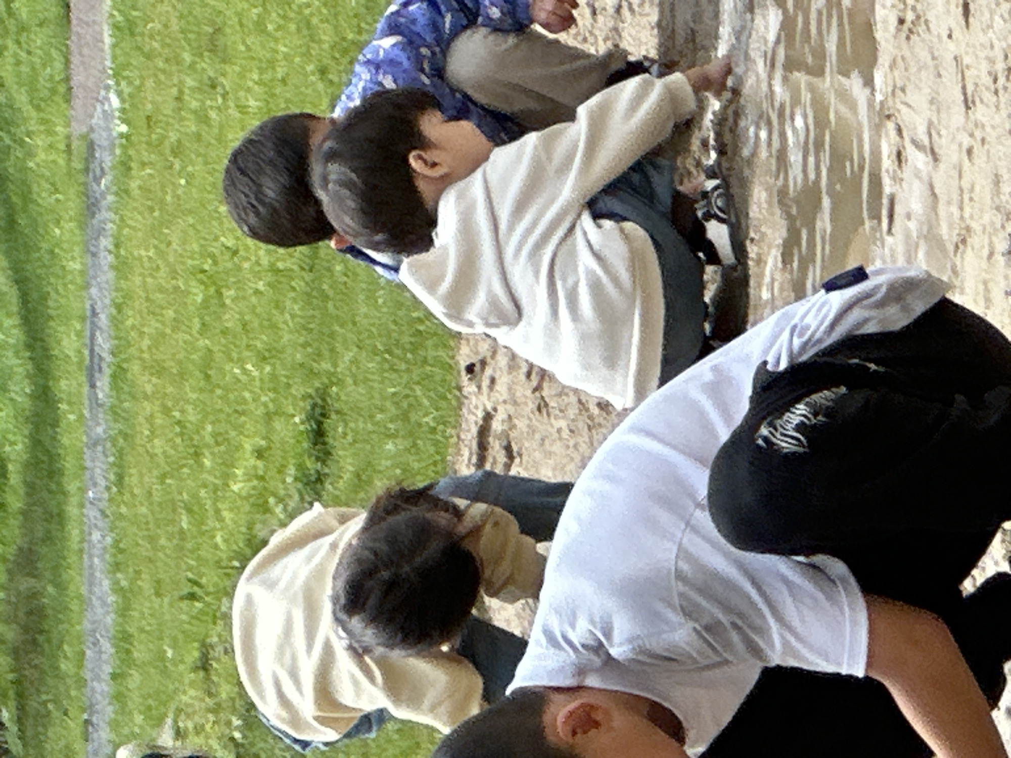 The image depicts a group of four young children outdoors in what appears to be a park or playground environment. The ground is a mix of grass and dirt, with a shallow puddle of water in the foreground. The children are dressed in casual clothing.
In the foreground, a child wearing a white t-shirt and black pants is partially visible, facing away from the camera and towards the puddle. To their right, another child in a light-colored hooded sweatshirt and dark pants is kneeling and reaching towards the puddle. Behind them, a child wearing a tan jacket and blue jeans is also bent over, appearing to interact with the ground. Further back, a child in a blue patterned jacket and tan pants is crouched down. The overall impression is of children engaged in outdoor play.
The location is identified as Znamya Oktyabrya, Russia. The environment suggests a mild weather day, suitable for outdoor activities.