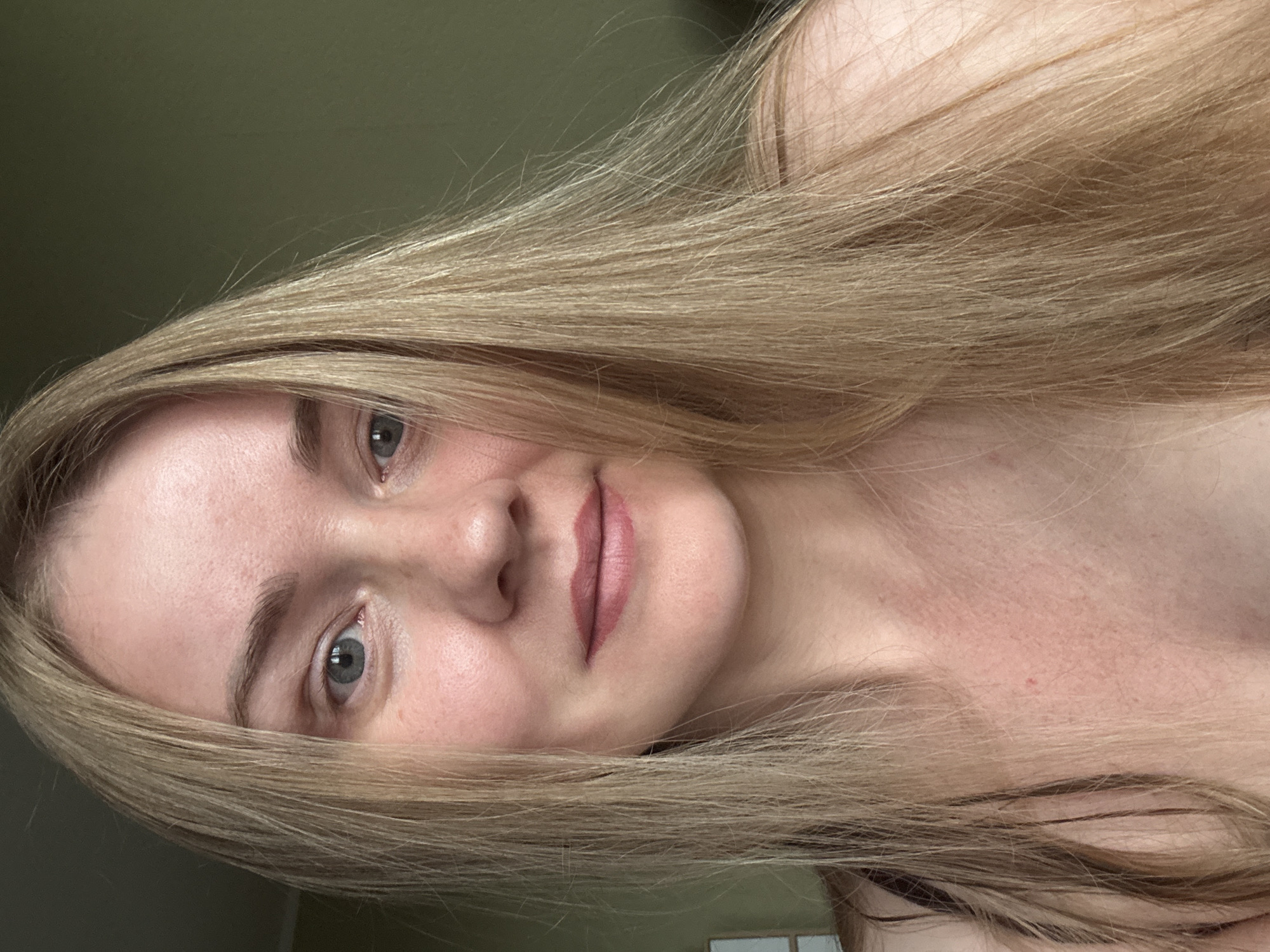 A close-up, front-facing selfie of a young adult female with long, straight blonde hair and light skin. She has blue-gray eyes, visible eyebrows, and her lips are lightly colored with lipstick. Her expression is neutral. She is wearing a dark-colored top, the straps of which are visible. The background is a plain, muted green wall. There is no identifiable information about the location within the city of Chaikovsky, Russia, nor any specific objects or actions occurring.