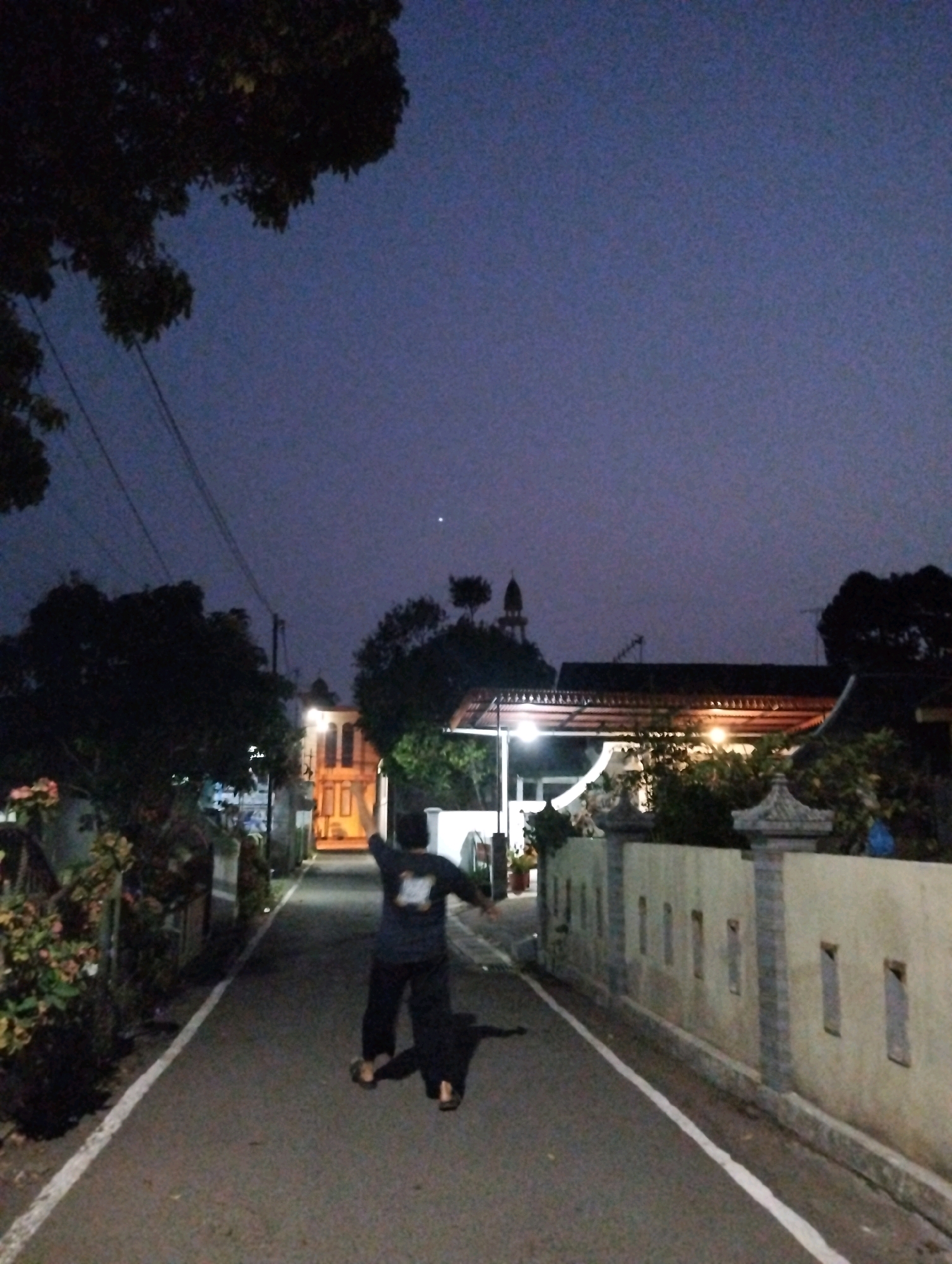 A low-angle, full shot captures an unidentified person in mid-jump on a paved road at dusk in Salatiga, Indonesia. The person, dressed in dark clothing and sandals, is facing away from the camera with arms outstretched. The road, lined with a low, cream-colored wall on the right and greenery on the left, leads to a building with illuminated windows in the distance. The sky is a deep purple, suggesting the transition from day to night. The scene is situated in a residential area, with houses and trees visible. The overall atmosphere is peaceful and serene.