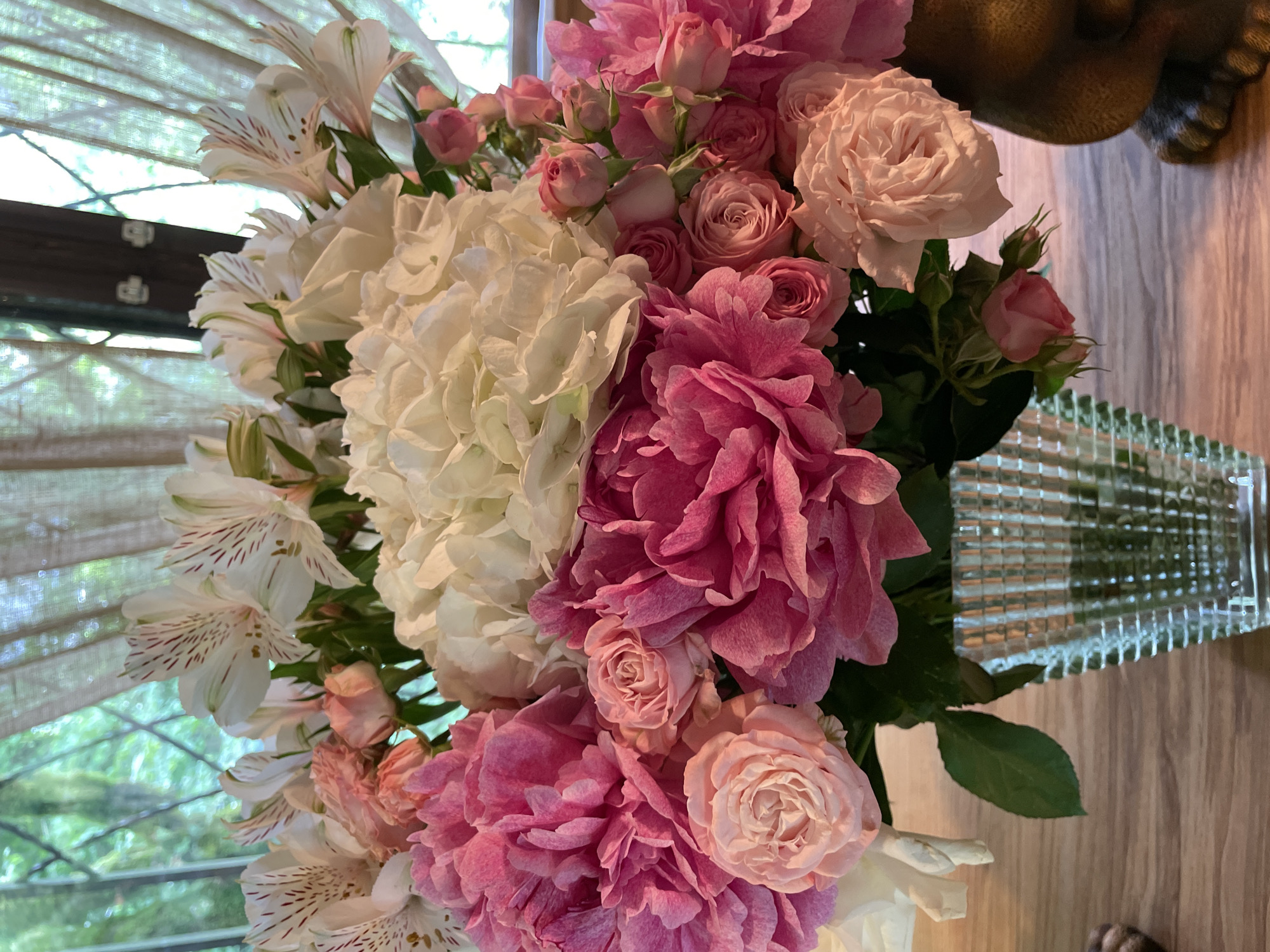A close-up shot shows a bouquet of flowers in a textured glass vase, sitting on a light-colored wooden surface. The bouquet contains white hydrangeas, pink peonies, various shades of pink roses, and white alstroemeria with reddish-brown speckles on the petals. The flowers are densely packed, creating a lush arrangement. A portion of a dark-colored, possibly bronze, object is visible in the upper right corner. In the background, a window with a light-colored curtain is partially visible. The photograph appears to be taken indoors. Location is not provided in the original image.