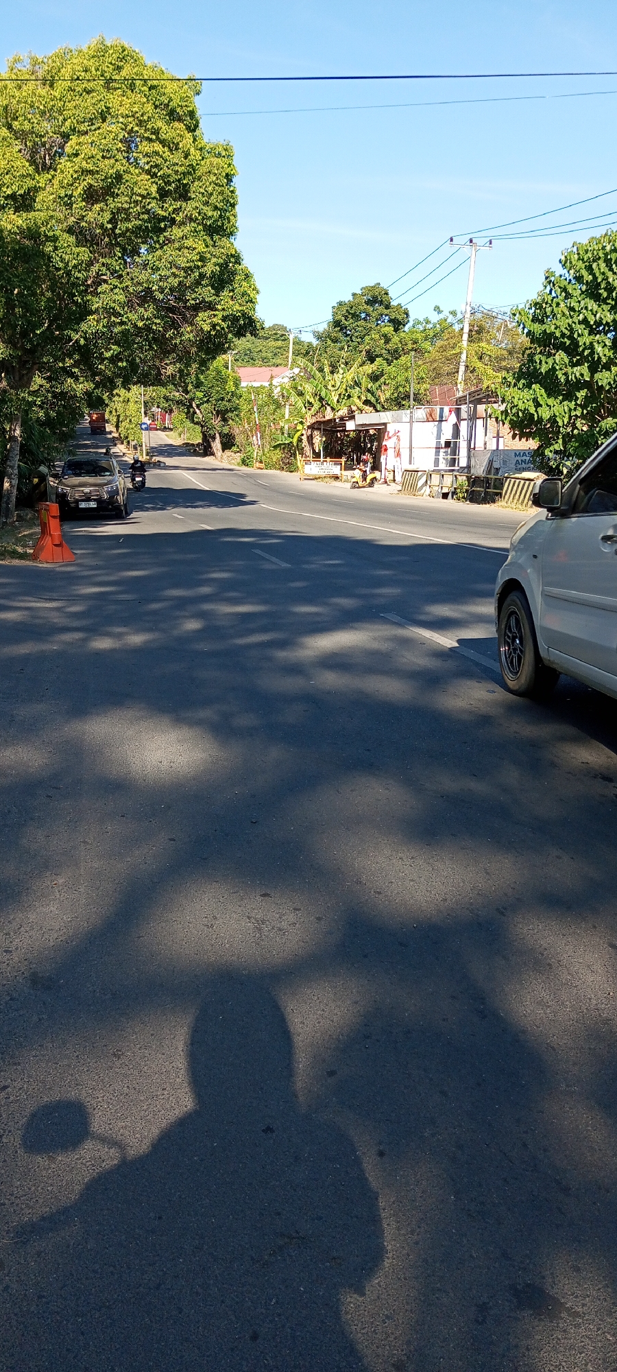 The image depicts a street scene in Bulupabbulu, Indonesia. The road is paved and stretches into the distance, lined with trees and vegetation. A white car is parked on the right side of the road, partially obscuring the view of the pavement. On the left side, a dark SUV is parked, with an orange traffic cone in front of it. A motorcycle is visible in the distance, on the left side of the road, heading away from the camera. There are buildings and stalls along the right side of the road, some with awnings. The sky is clear and blue, indicating a sunny day. Shadows from trees are cast across the road. The overall impression is of a rural or semi-urban setting in Indonesia during daylight hours.