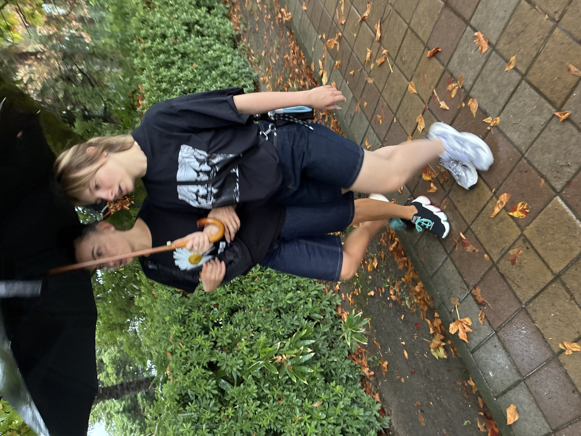 Two individuals, appearing to be an adult and a teenager, walk side-by-side under a black umbrella on a wet, tiled sidewalk lined with green foliage and fallen autumn leaves. The person on the left, likely the adult, wears a black t-shirt with a white daisy graphic and dark denim shorts, and carries a black bag. The person on the right, the teenager, also wears a black t-shirt with a graphic print and dark denim shorts. Both are wearing athletic shoes and walking to the right of the frame, as indicated by their leg positioning and the direction of their gaze. The environment suggests an outdoor setting in a city like Sochi, Russia, during an overcast or rainy day, with a paved walkway and dense greenery. The fallen leaves on the ground and the wet appearance of the path indicate autumn conditions.