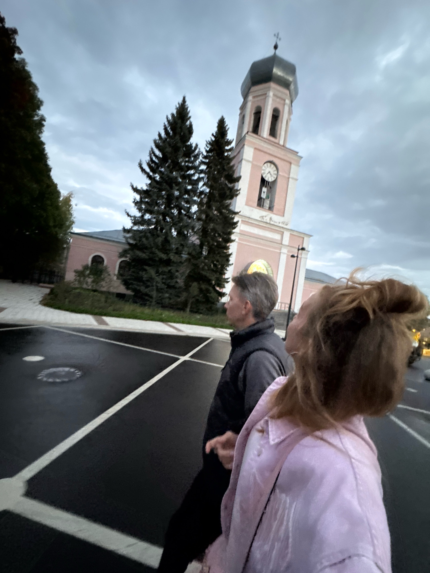 A medium shot captures a man and a woman standing on a wet, asphalt road marked with white lines, possibly a pedestrian crossing. The man, seen from the side in the foreground, wears a dark vest over a grey shirt. The woman, with her back to the camera, is in a light pink jacket. Behind them, a light pink building with arched windows is visible, partially obscured by dark green coniferous trees. Dominating the background is a tall, light pink clock tower with a dark, octagonal roof topped with a cross. The sky is overcast with grey clouds. The scene appears to be in Valday, Russia.