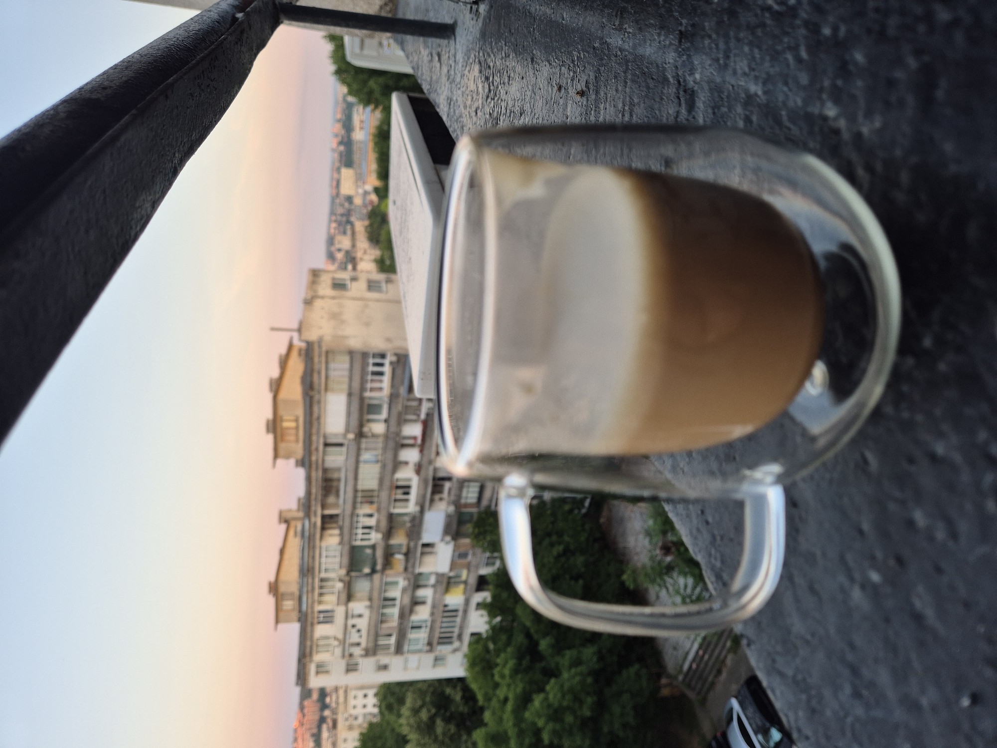 A close-up shot features a glass mug with a handle, filled with a light brown liquid, possibly coffee with milk. The mug is positioned on a dark, textured surface, likely a balcony railing, with the foreground out of focus. In the background, a city skyline is visible, characterized by apartment buildings of varying heights and architectural styles, some appearing aged. Lush green trees are interspersed among the buildings. The sky displays a gradient of colors, transitioning from a pale blue at the top to soft pink and orange hues near the horizon, suggesting either sunrise or sunset. A black railing runs across the top of the frame, partially obscuring the view.