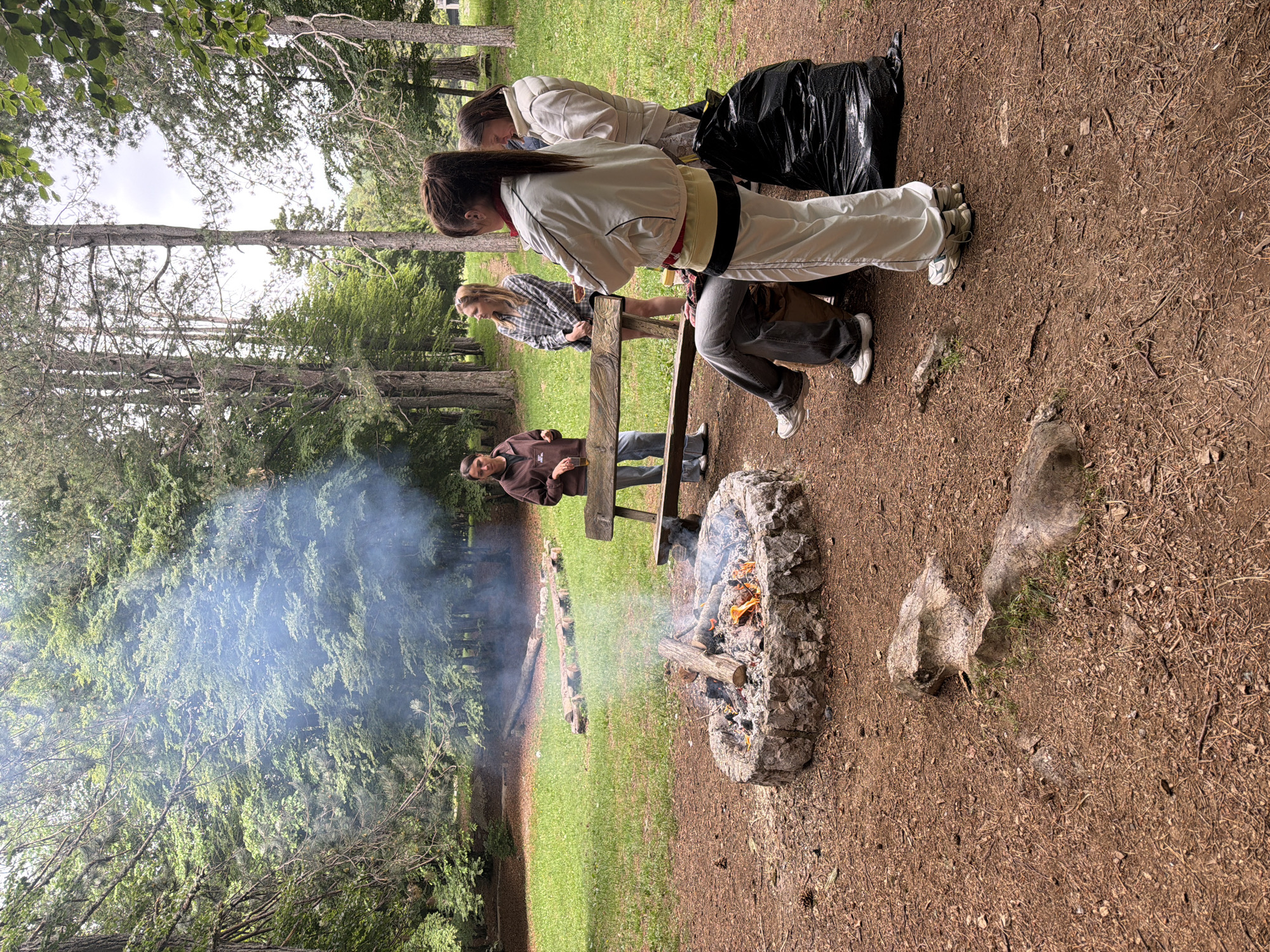 This image depicts an outdoor gathering around a campfire in a wooded area, identified as Mali Bostur, Montenegro. The scene is dominated by a stone fire pit located in the lower right, with a fire burning brightly and emitting a significant amount of smoke that drifts towards the left, partially obscuring the distant trees.
Several individuals are present, gathered around a wooden bench or table. One person in a white jacket and light-colored pants stands with their back largely to the viewer, leaning forward as if observing the fire or activity on the table. Behind them, another person in a lighter-colored jacket is partially visible. To the left, a person wearing a plaid shirt leans against the wooden structure, and further left, an individual in a dark brown hoodie stands near the bench, looking towards the fire. A full black trash bag rests on the ground near the group.
The setting is a natural environment with numerous tall trees and dense green foliage, suggesting a forest or park. The ground is a mix of brown earth covered with pine needles and patches of green grass. The lighting indicates it is daytime, likely under an overcast sky, providing diffused light and no harsh shadows. The overall impression is one of a relaxed, social occasion centered around a campfire. There is no visible text in the image.