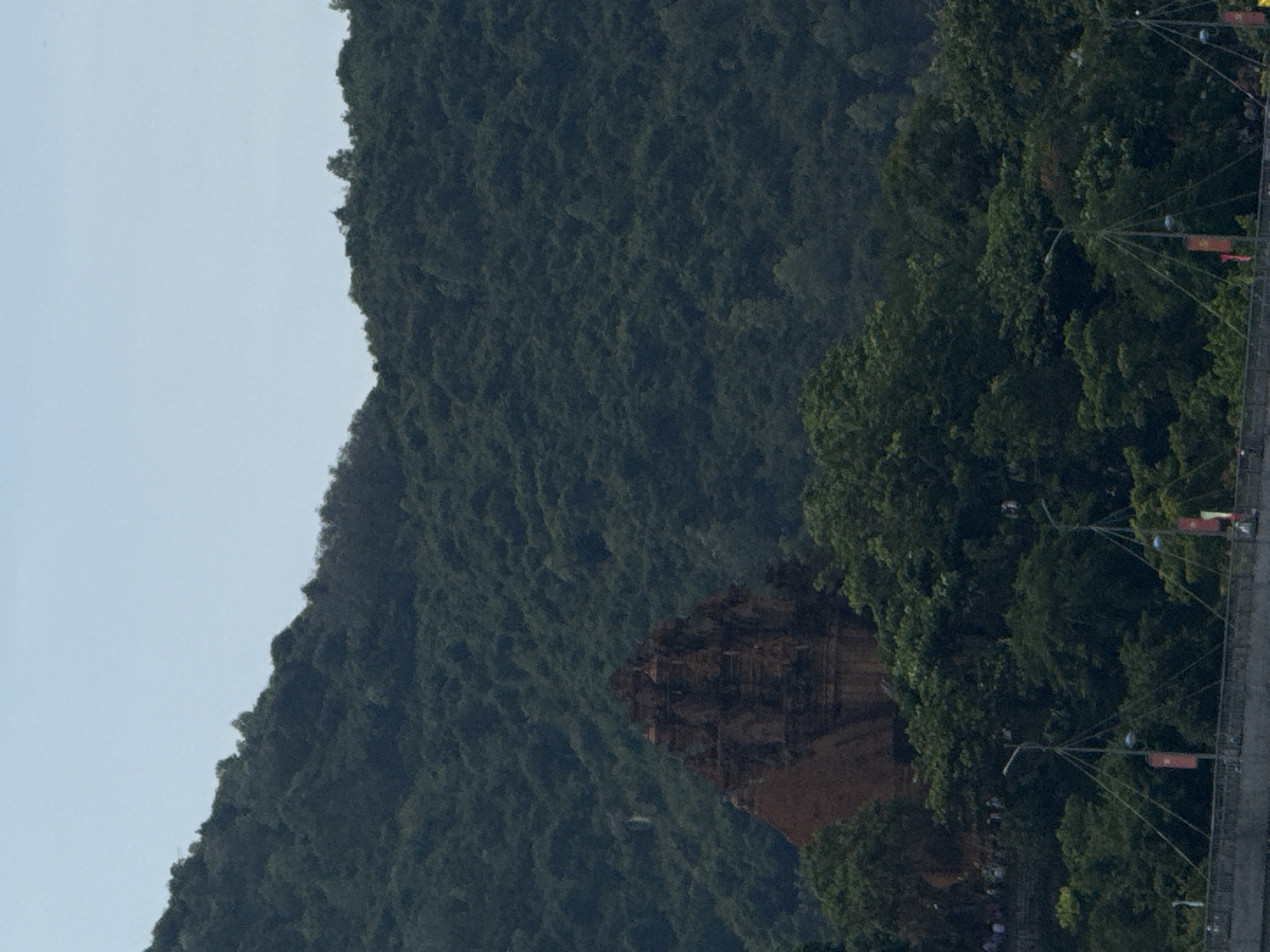 A slightly tilted medium shot shows the Po Nagar Cham Towers in Nha Trang Ward, Vietnam.
The towers, constructed of dark brown brick, are partially obscured by dense green trees. A large, forested mountain dominates the left side of the frame, contrasting with the light blue sky. On the right, street lamps and flag poles line a path, indicating human activity. A few white-clad individuals are visible in a small cluster beneath the trees, but their actions are indiscernible.