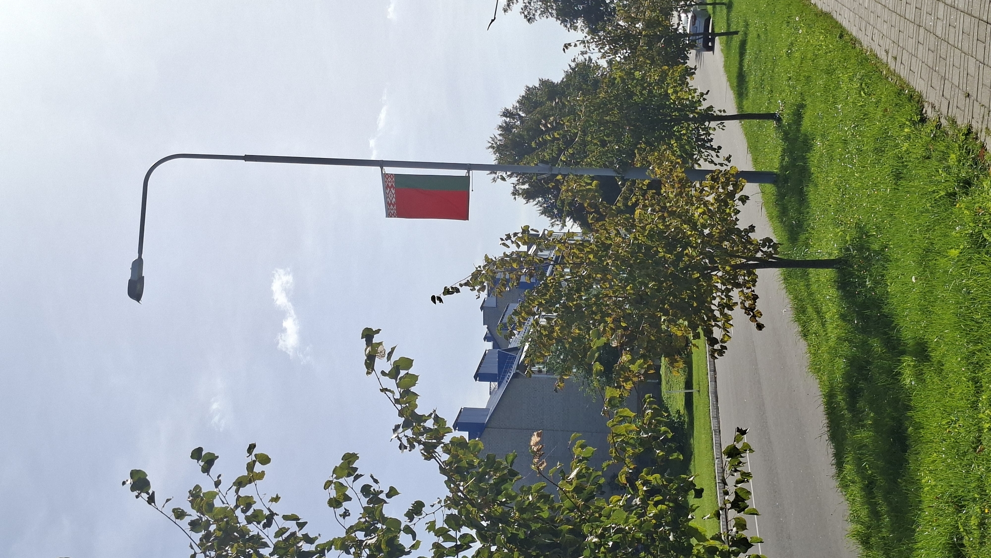The image depicts a street scene in Myadzyel, Belarus. A lamppost with a Belarusian flag hangs from it stands on the right side of the frame. A road runs along the left, with a sidewalk on the right. Trees line the sidewalk, and buildings are visible in the background. The sky is blue with some clouds, and the sun is shining. The overall scene is peaceful and ordinary, suggesting a typical day in the city.